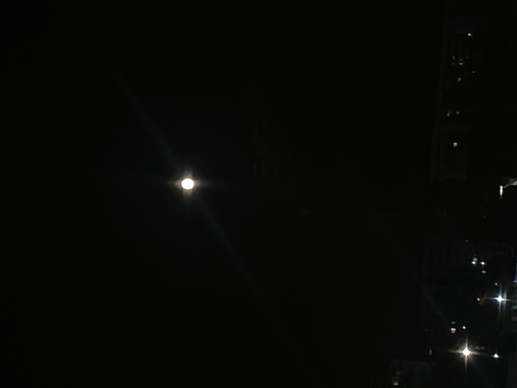 The image depicts a nighttime scene in Iași, Romania, dominated by a dark sky with a bright, full moon. The moon is positioned slightly off-center in the upper portion of the frame, casting a faint glow and a noticeable lens flare that extends diagonally downwards. Below the moon, the silhouette of a city skyline is visible. The skyline consists of dark, indistinct buildings, some of which have illuminated windows, suggesting residential or commercial structures. Streetlights are also visible as bright points of light, creating starburst effects due to the camera's exposure. The overall atmosphere is serene and quiet, characteristic of a city at night. No individuals are present in the image.