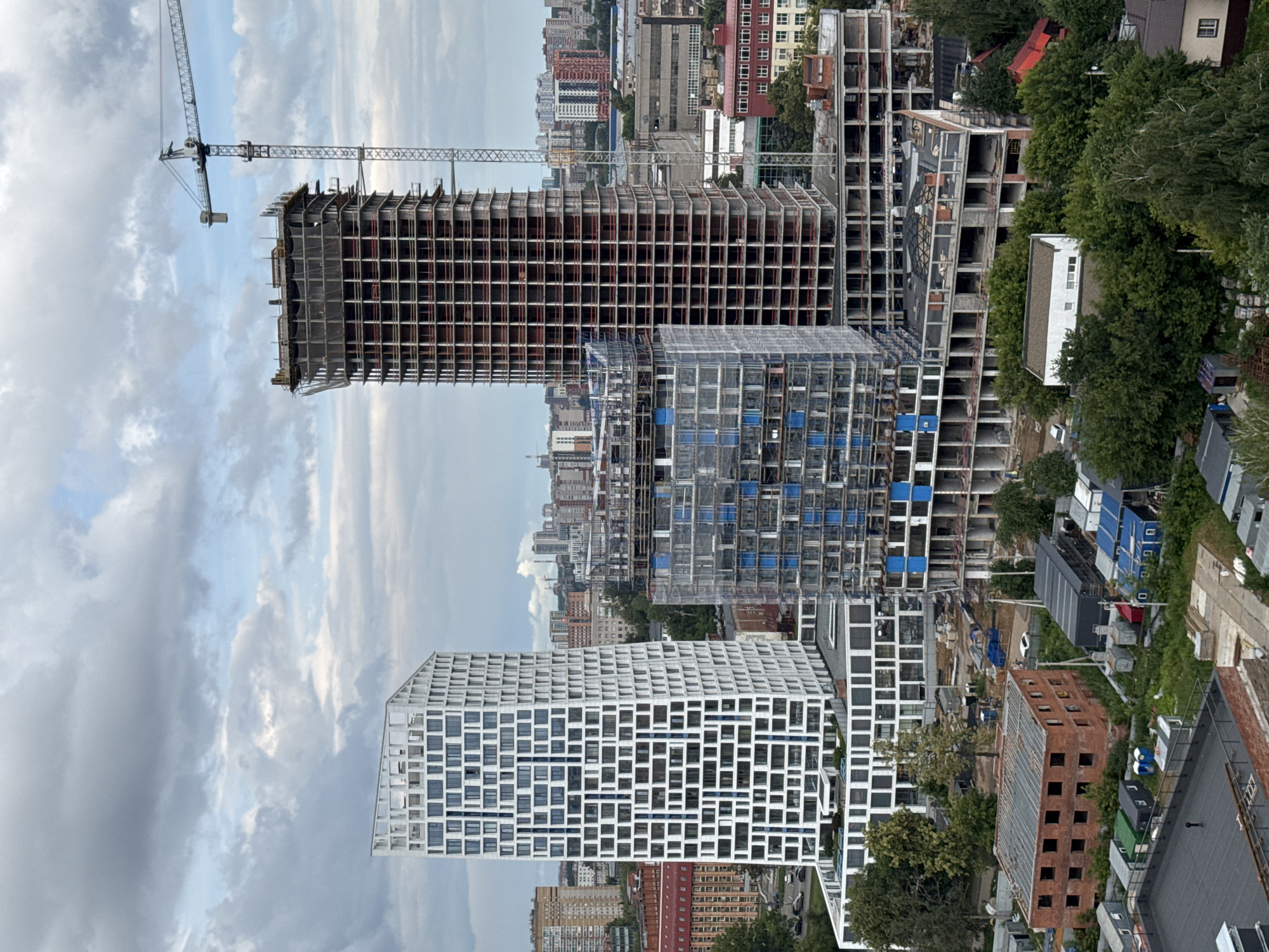 The image shows a construction site in Izhevsk, Russia. The main subjects are several unfinished buildings in various stages of construction, a large crane, and surrounding trees. The scene is an urban environment with multiple high-rise buildings visible in the background. The construction appears to be ongoing, with exposed frameworks and incomplete facades on some structures. One building is particularly striking with its white, grid-like exterior and numerous rectangular windows. Another partially built structure is made of red brick. The sky is cloudy, suggesting it may be daytime with overcast weather. No people are clearly visible in the image. There is no visible text. The overall impression is one of active urban development.