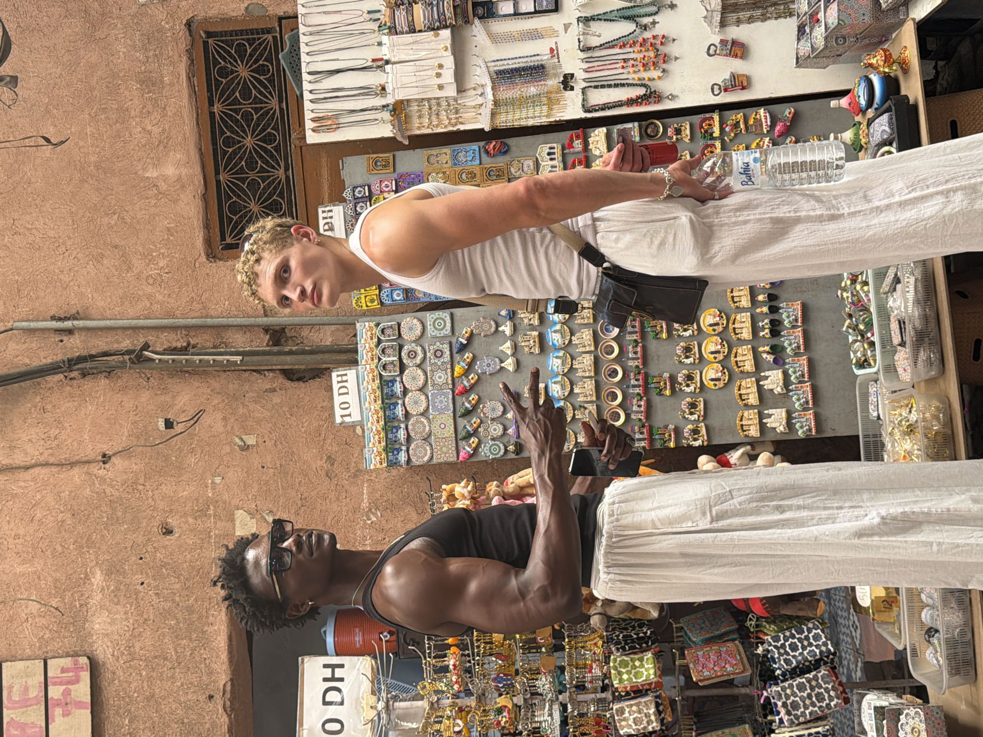 Two young men stand in front of a market stall in Marrakech, Morocco. The man on the left, with dark skin and short, dark, curly hair, wears sunglasses, a black tank top, and loose white pants. He gestures with his right hand, holding a phone in his left. The man on the right, with light skin and short, blonde, curly hair, wears a white tank top and loose white pants. He holds a bottle of "Bahia" water in his right hand and has a dark shoulder bag. The stall behind them is filled with numerous small souvenirs, including magnets, keychains, and jewelry, displayed on boards and shelves. The wall behind the stall is a textured, reddish-brown color, typical of many buildings in Marrakech's medina. The overall atmosphere suggests a bustling tourist area.