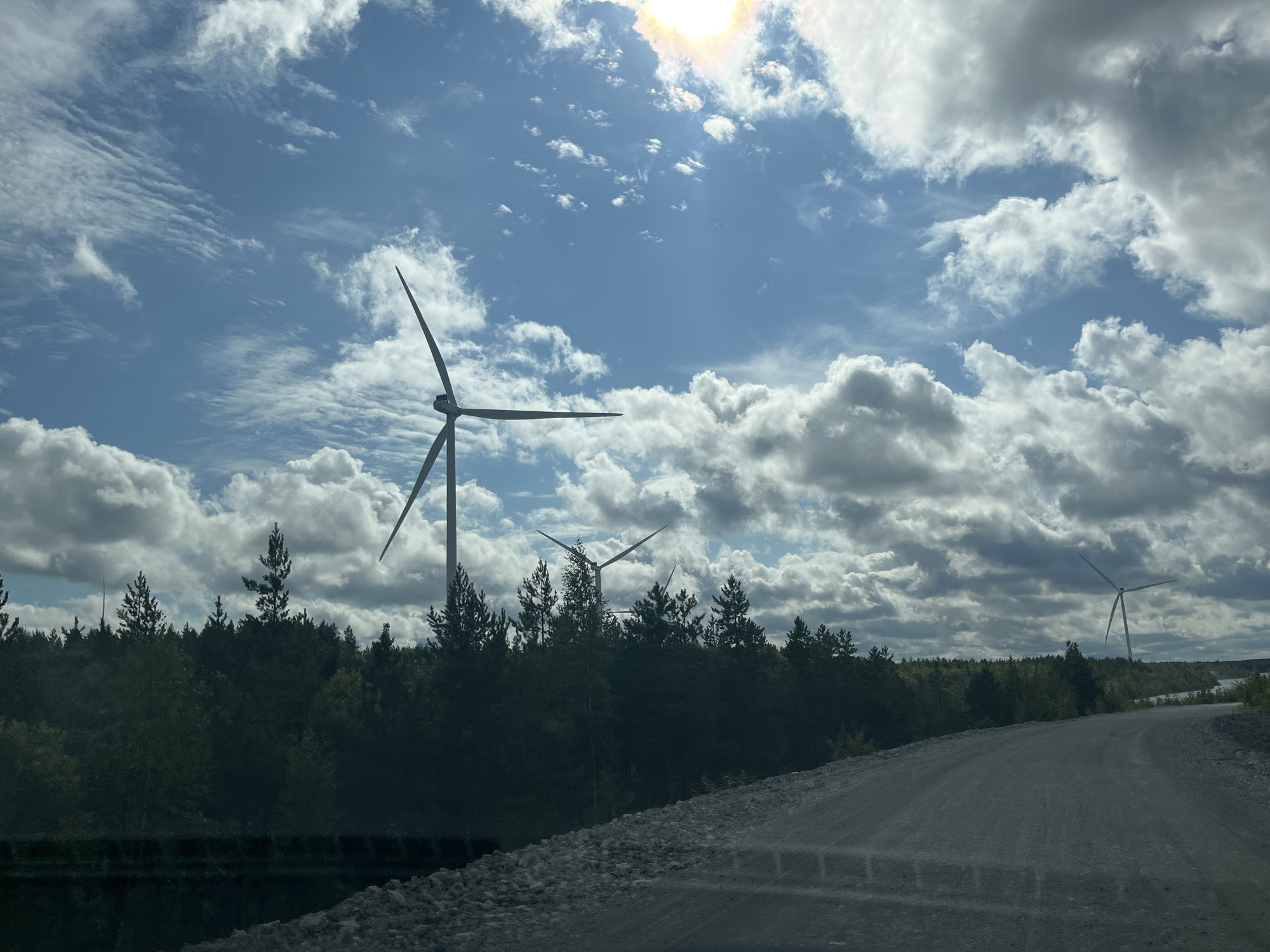 The image depicts a landscape scene in Aidu, Estonia, featuring several wind turbines set against a backdrop of a bright, cloudy sky. In the foreground, a dirt road curves to the right, its surface appearing rough and rocky. Dense, dark green trees form a line along the horizon, partially obscuring the lower sections of the wind turbines. The largest wind turbine is prominent on the left side of the frame, with its blades extended. Two other turbines are visible in the mid-ground and background, also silhouetted against the sky. The sky itself is a vibrant blue, populated by a mix of white and gray clouds, with the sun shining through, creating a slight halo effect. The overall impression is one of a rural, windswept environment, likely a wind farm. There are no people or animals visible in the image.