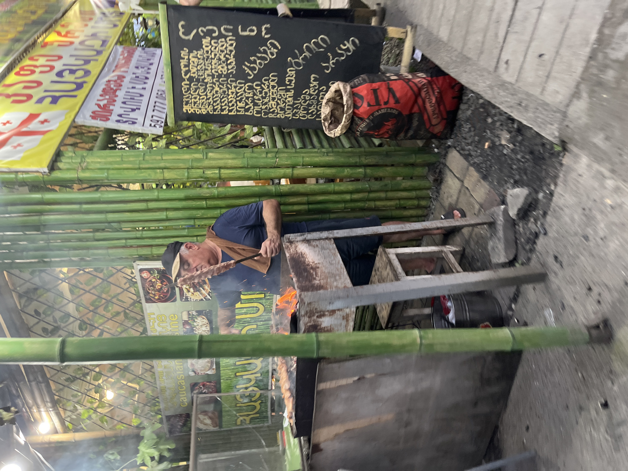 In Ureki, Georgia, a man stands in front of an outdoor food stall, tending to a grill laden with meat skewers. He is wearing a dark blue t-shirt, dark shorts, and a black baseball cap. A brown towel is draped over his shoulder. The grill is positioned over a fire, with flames visible. Behind the man, a wall constructed of vertical bamboo poles supports a green lattice structure, possibly a canopy. Several signs are displayed, including one with Georgian script and flags, and another with the words "Caucasian Cuisine." A blackboard menu also features Georgian text. A large red and black sack, presumably containing charcoal, rests on the ground near the stall. The area around the stall is paved with interlocking stones and scattered charcoal. The overall impression is of a casual, open-air dining establishment.