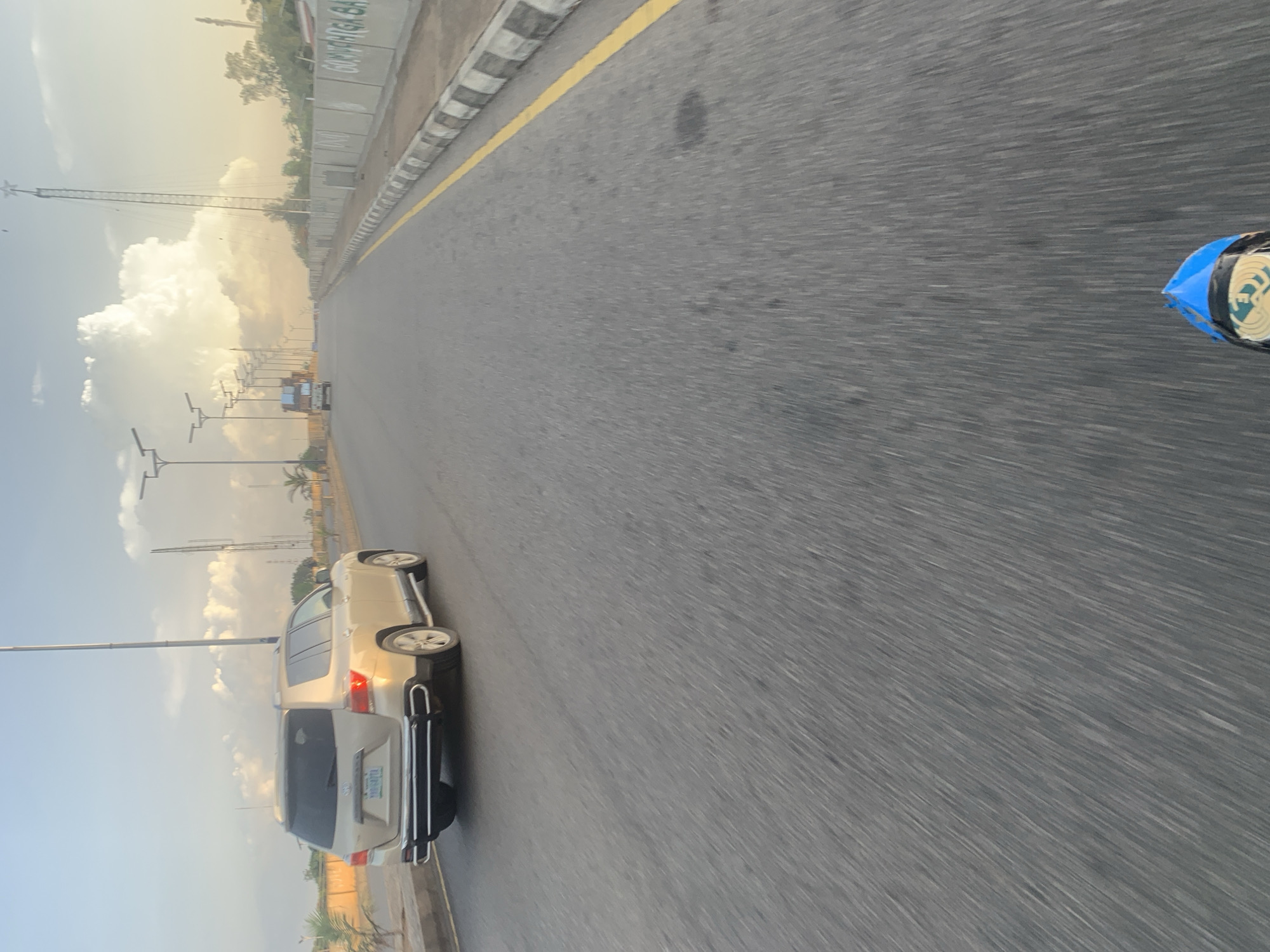 The image depicts a wide, paved road in an urban environment, likely in Jos, Nigeria, under a sky with scattered clouds and warm, diffused sunlight. A tan SUV is in the left lane, seen from its rear. To the right of the road, a concrete barrier with black and white markings lines the pavement. Further to the right, a truck is visible in the distance on the same road. Along the right side of the road, utility poles with lights are spaced out. In the background, there are buildings and trees, with a tower-like structure featuring a star at its apex. The road surface shows motion blur, indicating movement. A small portion of what appears to be a blue and black object, possibly part of a vehicle or equipment, is visible at the very bottom of the frame. The overall scene suggests an outdoor, daytime setting on a thoroughfare.