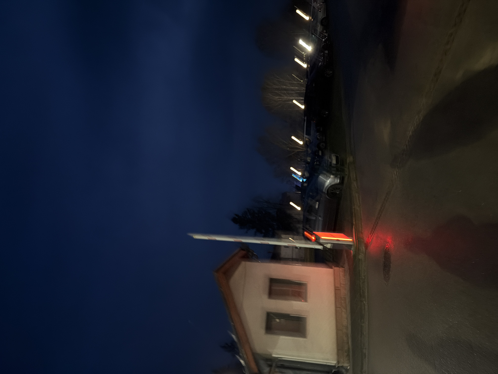 At night, a building with a boom barrier is positioned near a street lit by street lights. The building is located at the entrance of the Opticoelectron industrial park in Panagyurishte, Bulgaria. The barrier is visible. Cars are parked on the side of the street behind the gate. The sky is dark blue.