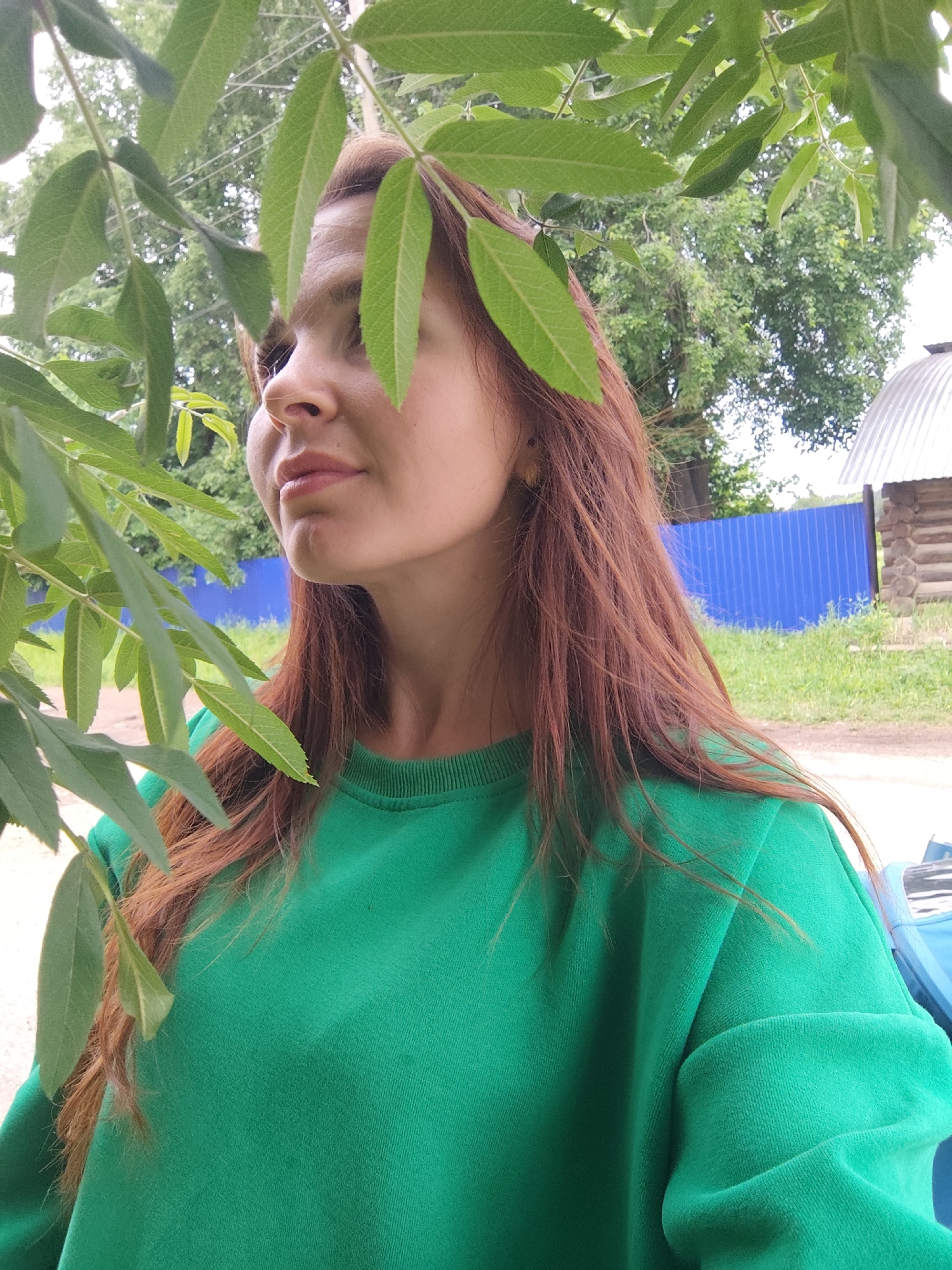A woman with red hair is standing outdoors in the city of Фоки, Russia. She is wearing a green sweatshirt and is partially obscured by green foliage. In the background is a blue fence, green grass, and a small wooden structure with a metal roof. The woman is looking off to the side.