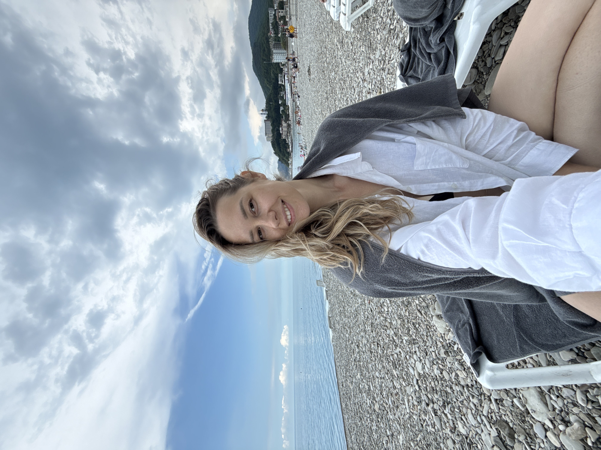 A woman with blonde, wavy hair is taking a selfie on a pebbled beach in Nebug, Russia. She is wearing a white unbuttoned shirt over a black bikini and a dark grey towel is draped around her shoulders. She is sitting on a white lounge chair. The ocean is visible behind her, and the sky is cloudy with patches of blue. In the background, there are more lounge chairs, people, and buildings on a hillside.