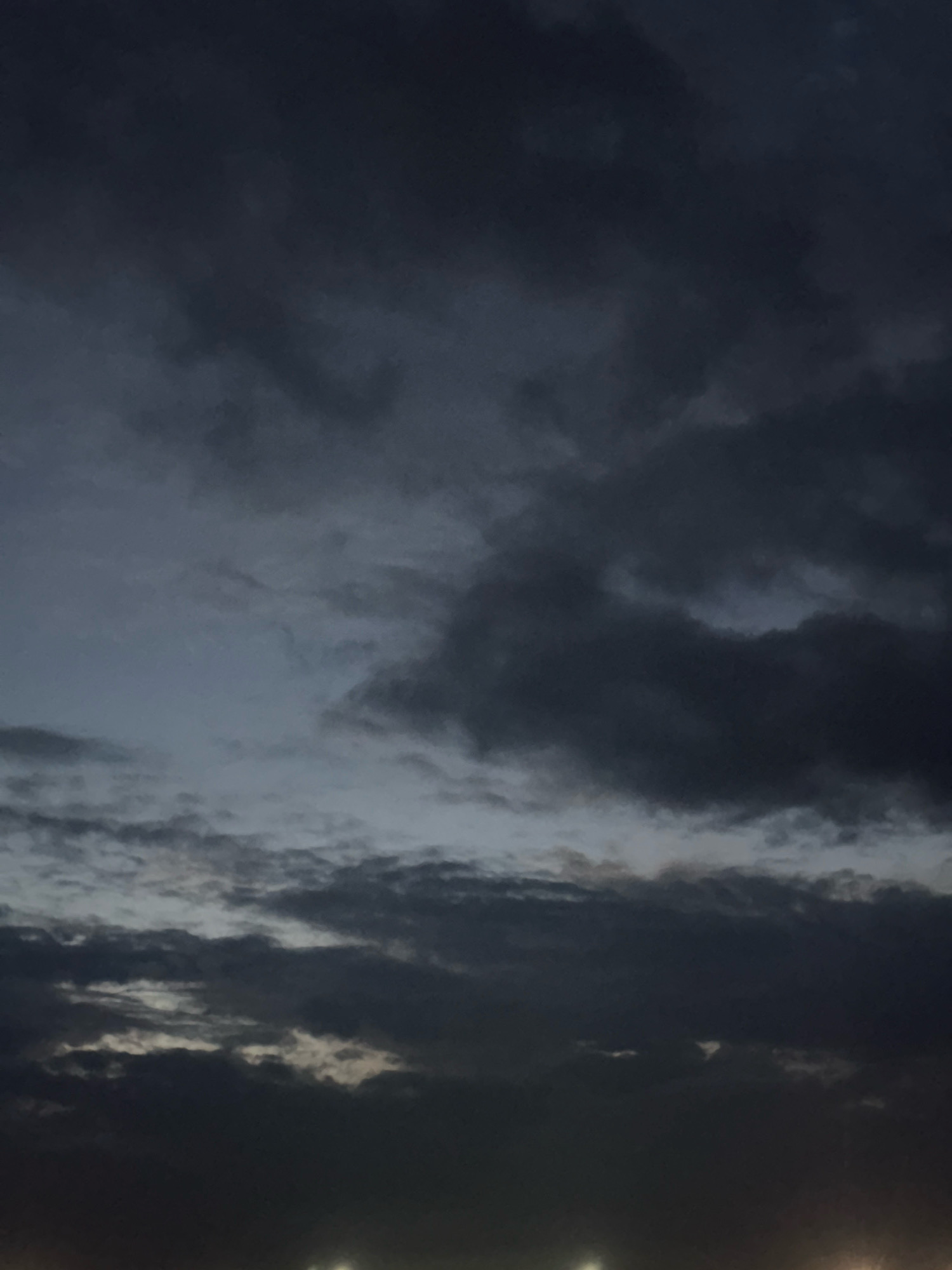 The image depicts a dark, overcast sky with numerous clouds. The lighting suggests it is either dusk or pre-dawn, with a hint of light near the horizon. There are no visible people, subjects, or specific objects that can be identified. The environment appears to be an open area, possibly a field or a body of water, given the limited visible details at the bottom of the frame which show faint lights that could be from distant streetlights or structures. No specific location within Galinė, Lithuania can be identified from the image itself, as it solely focuses on the sky.