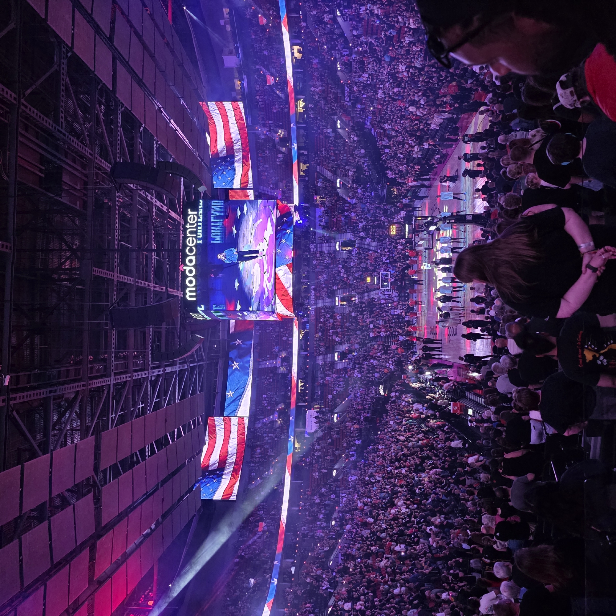 The image depicts a large indoor arena, identified by the "Moda Center" sign. The arena is filled with a diverse crowd of people, many of whom appear to be spectators, occupying tiered seating that surrounds a central court area. The court is lit and shows a number of individuals, likely performers or participants, in various poses, some standing and others in motion. Large American flags are prominently displayed, hanging from the ceiling and incorporated into the visual presentation on the large screens. The overall atmosphere suggests a public event, possibly a sporting match or a concert, given the scale of the gathering and the elaborate lighting. The color palette is dominated by purples and reds from the lighting, with the blue and white of the flags providing a contrasting element. The image is taken from a vantage point within the seating area, looking down towards the court and the stage.