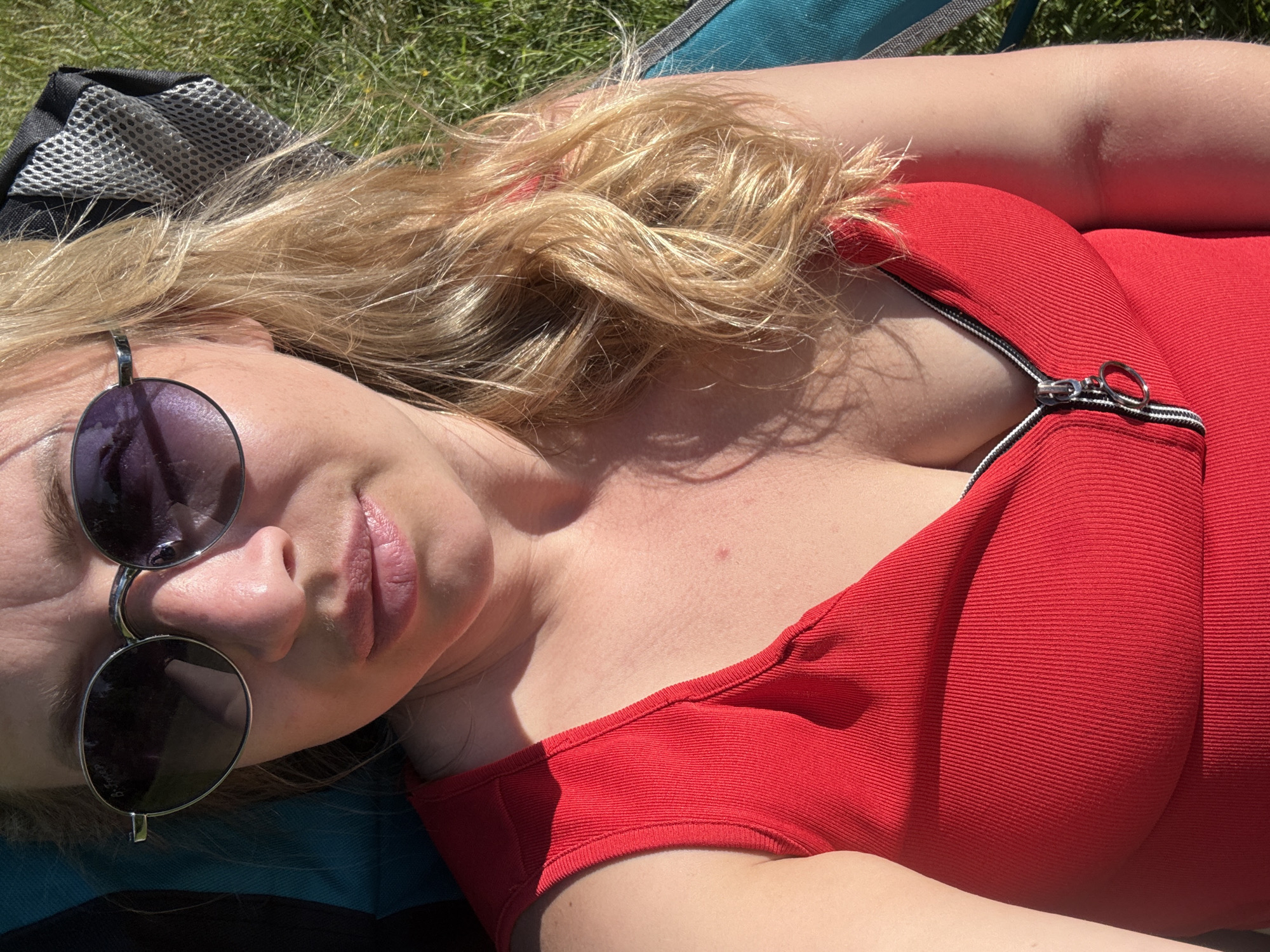 An overhead, medium-close shot shows a fair-skinned woman with blonde hair and sunglasses lying in a chair in a grassy area. The woman is wearing a red, ribbed tank top with a zipper that is partially unzipped. The background is blurred green grass and a partial view of a gray mesh headrest on a chair. The lighting suggests it is daytime and sunny, with the sun casting shadows on the woman's skin and hair. The overall scene appears to be outdoors, possibly in a park or backyard.