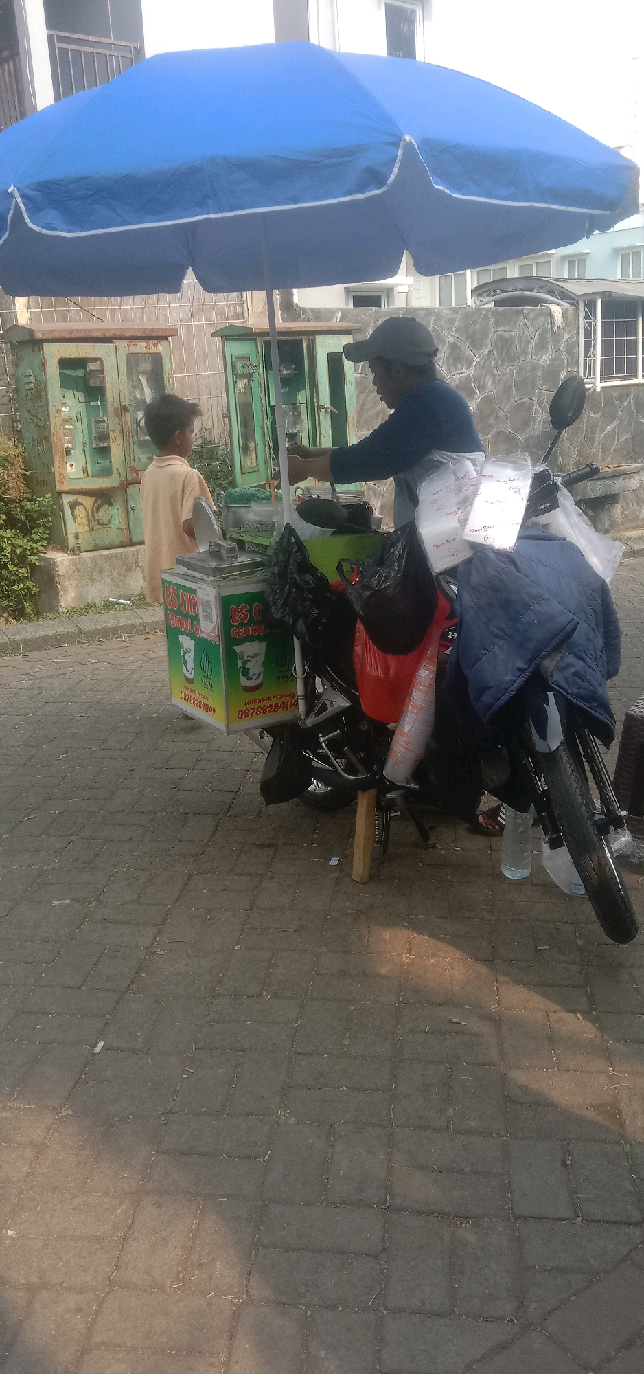 In Sukanagalih, Indonesia, a street vendor, a man wearing a dark blue shirt and a baseball cap, stands behind a motorcycle-mounted cart. The cart is adorned with a large blue umbrella and displays the text "ES CINCIN" and a phone number, "087882841149". A young boy in a light orange shirt stands to the left of the cart, facing away from the camera. The scene is set on a paved walkway, with green utility boxes visible in the background. The lighting suggests it is daytime, possibly late afternoon given the shadows.