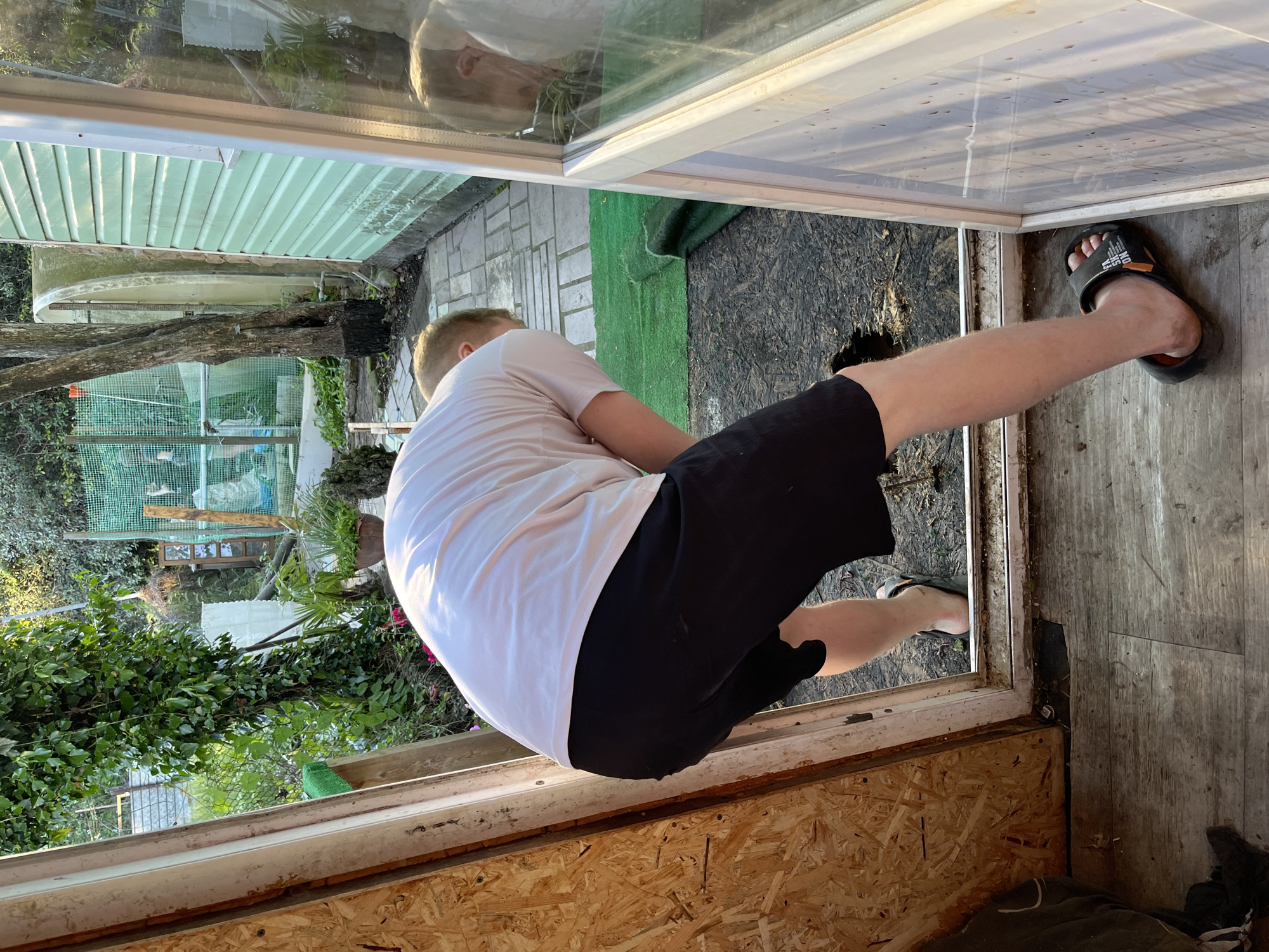 A man wearing a white t-shirt and black shorts is squatting in a doorway. He is wearing sandals on his feet. The doorway leads to an outdoor area with greenery, including plants and trees. The doorway frame appears to be made of wood and possibly includes some particleboard. The interior floor of the doorway is made of wood. The scene is likely outdoors and has an informal, possibly domestic setting. There is no indication that the image was taken in Vesyoloye, Russia.