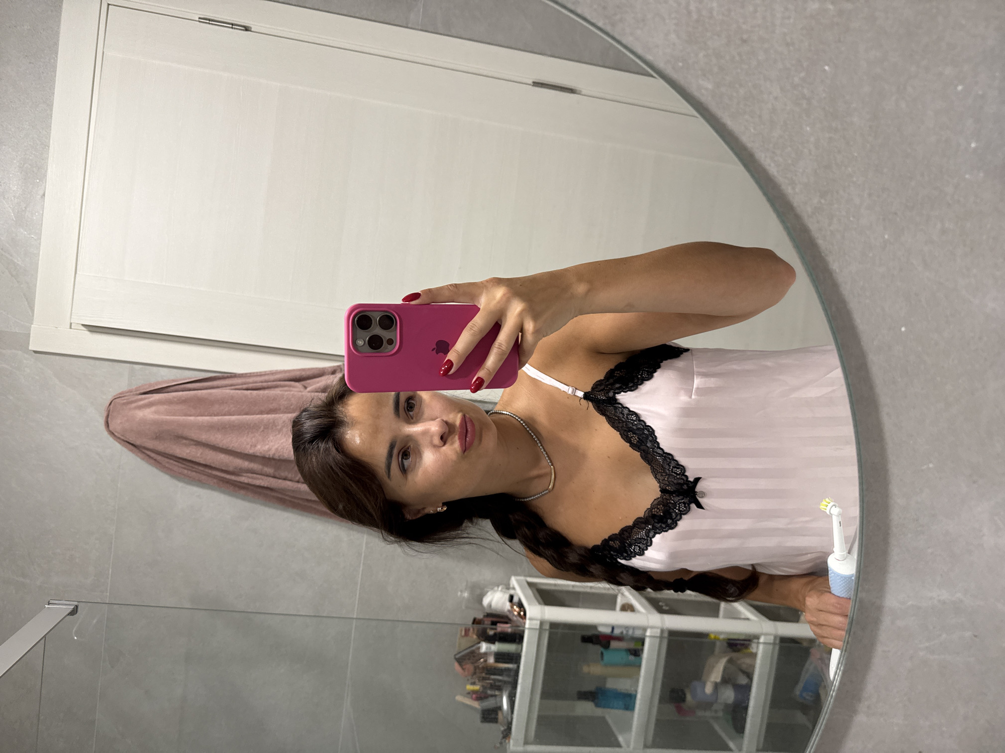 A woman with brown hair styled in a braid, wearing a pink camisole with black lace trim, stands in a bathroom and takes a selfie in a mirror. She is holding a pink smartphone in her right hand and an electric toothbrush in her left hand, which is resting on the edge of the sink. The bathroom features grey tiled walls, a pink towel draped over a shower rod, and a white door with vertical paneling. A white shelving unit with various toiletries is visible to the left of the woman.