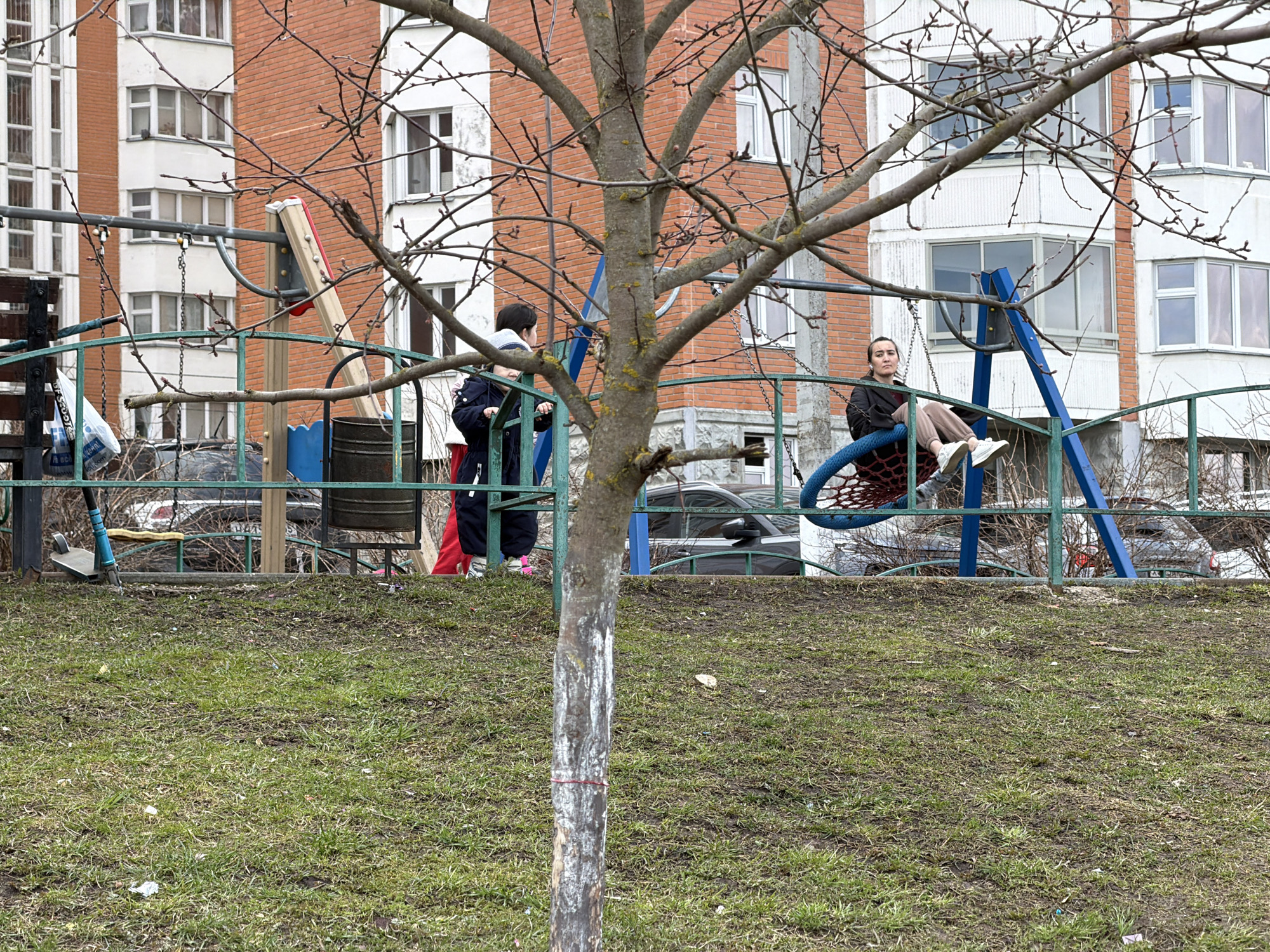 The image depicts a scene in Brekhovo, Russia. In the foreground is a young tree, its trunk painted white, with bare branches. Beyond the tree, a playground is situated on a patch of grass that shows signs of wear. A child stands near the playground equipment, appearing to look at something. A woman sits on a circular swing set. In the background, apartment buildings are visible, predominantly constructed of brick and white concrete, with numerous windows. Several cars are parked near the playground, partially obscured by the playground fence. The overall scene has a muted color palette, reflecting a cold weather, probably spring or autumn.