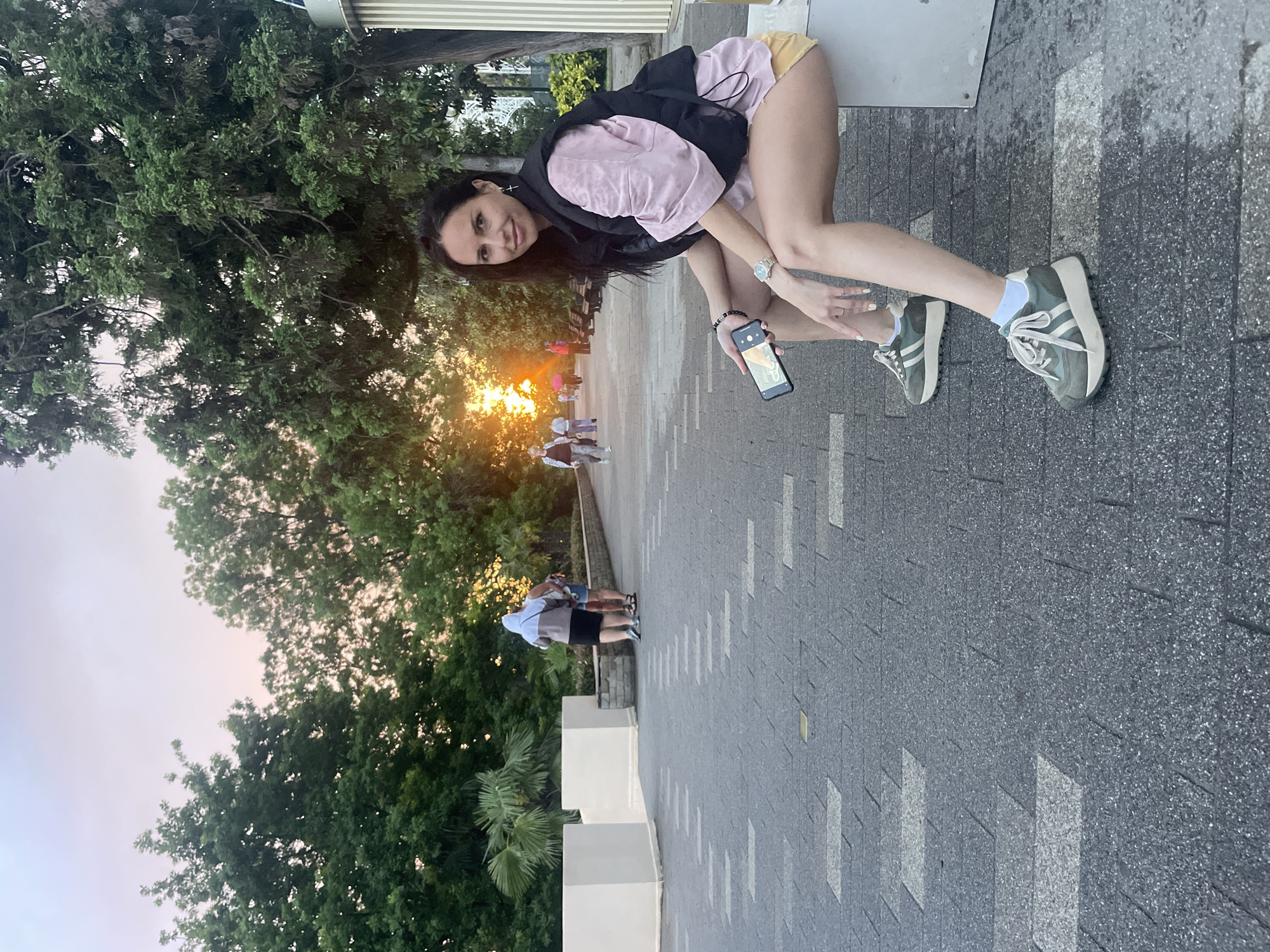 A woman with long dark hair, wearing a light pink top, a black vest, and shorts, is crouched on a stone-paved surface while holding a smartphone. She is angled towards the viewer, with a slight smile. Her feet are clad in green and white sneakers with white socks.
In the background, there is a pathway lined with trees and other vegetation. In the distance, figures of people can be seen along the pathway, with a bright light source, seemingly the sun, situated further down the path. The pathway is bordered by a low, white wall. The environment appears to be outdoors, possibly a park or public space.
Based on the context of the question, this location is in the city of Sochi, Russia.