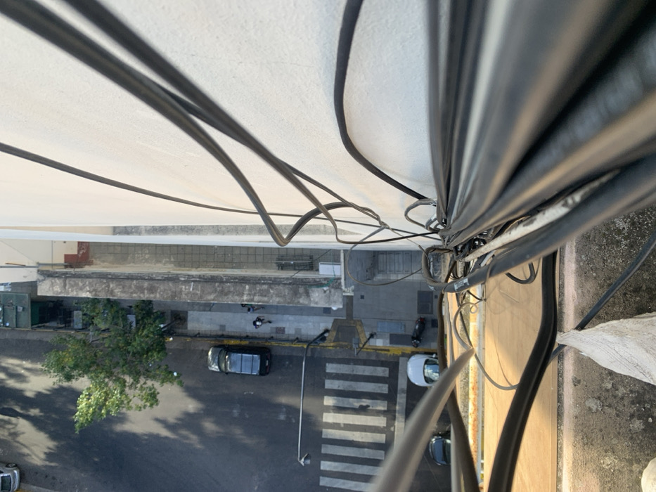 A high-angle shot shows a street scene from above, with several cars parked and a pedestrian crossing a crosswalk. A tree is visible on the left side of the street. Electrical cables dominate the foreground, creating a network of lines that obscure parts of the view. The perspective makes the scene appear somewhat distorted, emphasizing the height from which the photo was taken. Buildings are visible in the distance.