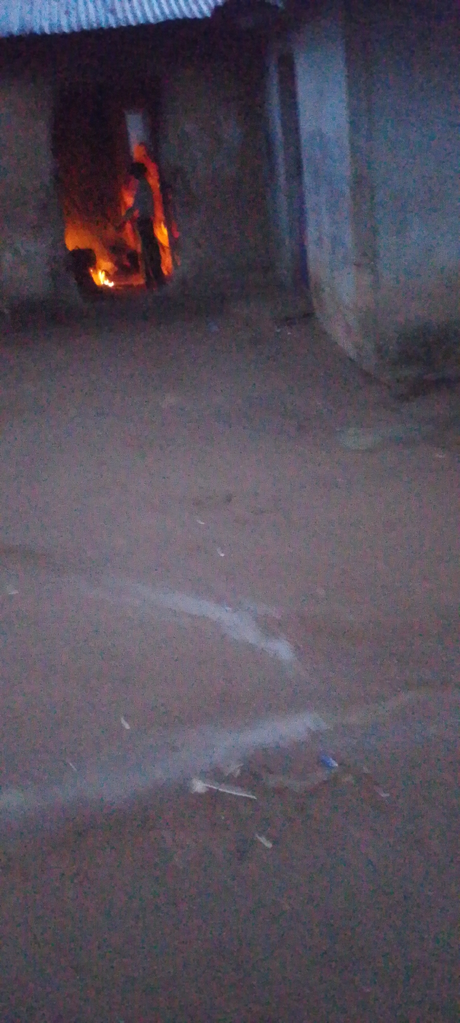 The image depicts a nighttime scene outside a dimly lit structure, possibly a dwelling or shelter, with an open doorway revealing a fire burning within. A silhouetted figure, appearing to be a person, is partially visible standing near the fire. The ground in front of the structure is dark and appears to be unpaved. There are faint, irregular white markings on the ground, possibly chalk or paint, forming abstract shapes. The overall lighting suggests it is either dusk or nighttime, with the fire being the primary light source within the structure. The texture of the ground is rough, indicative of dirt or gravel. The structure itself has a corrugated metal roof visible at the top of the image. The scene is devoid of any clear weather indicators or specific location cues. The mood is somewhat ambiguous, with the fire and darkness potentially evoking feelings of warmth, danger, or mystery. There is no visible text in the image.