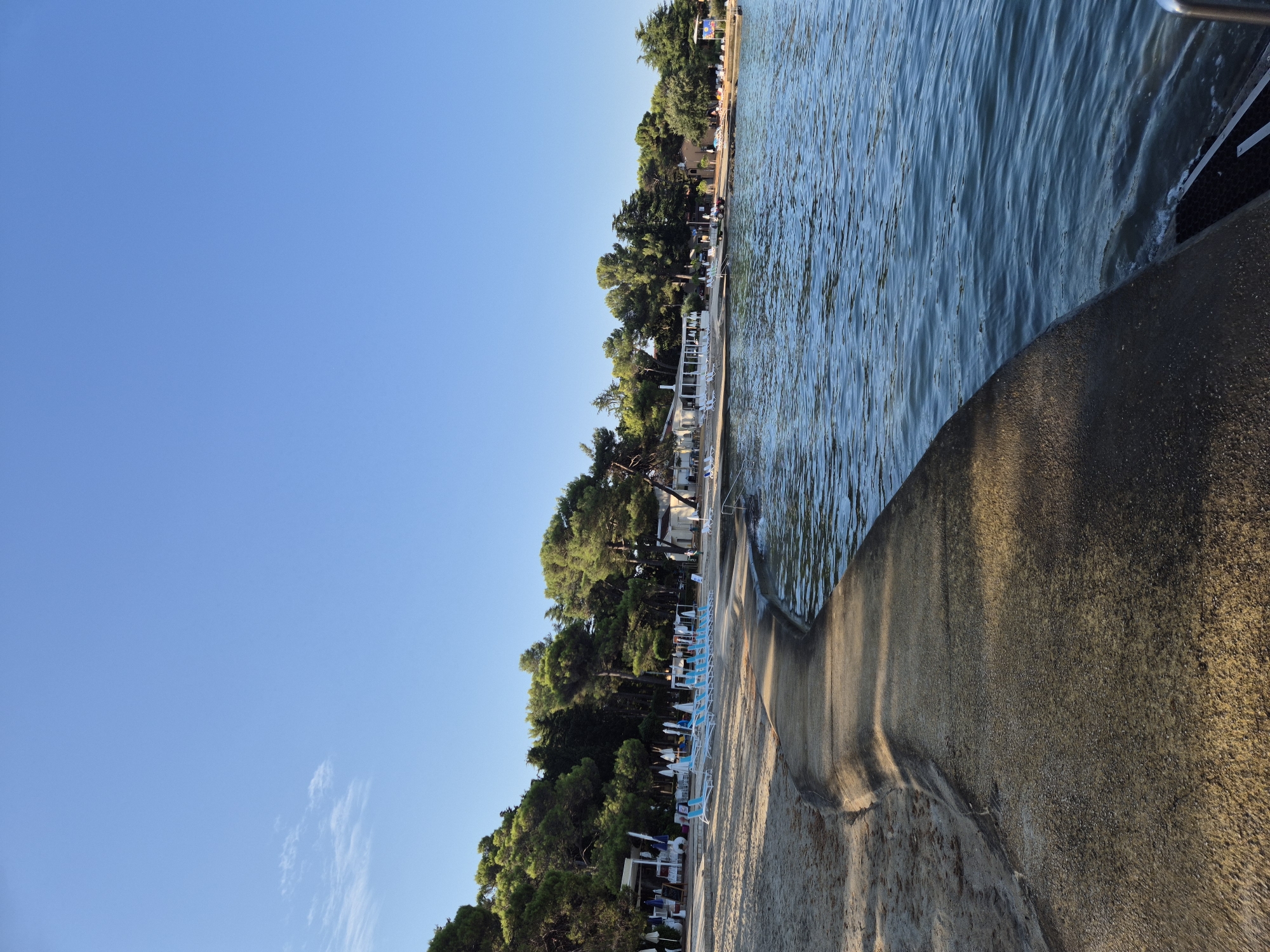 The image depicts a serene view of a beach resort on a clear, sunny day, likely in Monterol, Croatia. In the foreground, a curved concrete walkway separates the sandy beach from the calm, blue water. The beach itself is lined with rows of blue lounge chairs and white umbrellas, suggesting a well-maintained tourist area. Beyond the beach, a dense line of tall green trees forms a natural backdrop, with a few white buildings peeking through the foliage. These buildings appear to be part of the resort, possibly housing restaurants or accommodations. The sky is a vibrant, cloudless blue, indicating pleasant weather. There are no visible people or specific actions taking place in the scene. The overall impression is one of a peaceful and inviting seaside vacation spot.