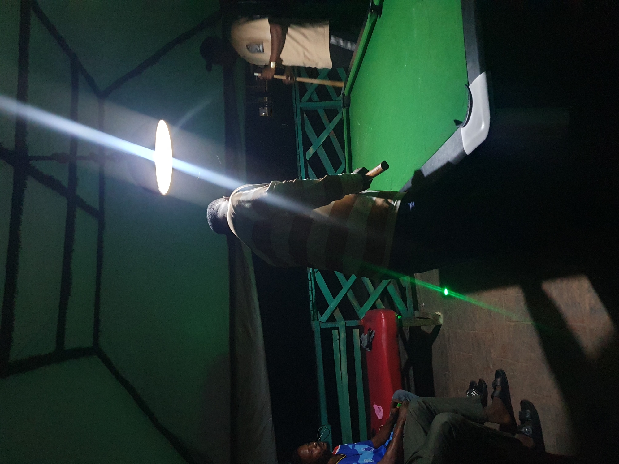 In Lokoja, Nigeria, a dimly lit, open-air pavilion features a green billiard table in the foreground. Two men are visible. One, wearing a tan shirt and black shorts, stands on the right, holding a pool cue. The other man, wearing a striped shirt, is further left, facing away from the camera, also holding a pool cue. A red bench with some fabric items on it is positioned behind the man in the striped shirt. The pavilion's structure, with dark beams against a green fabric ceiling, is visible above. The environment appears to be an outdoor recreational area, possibly a bar or club, at night.