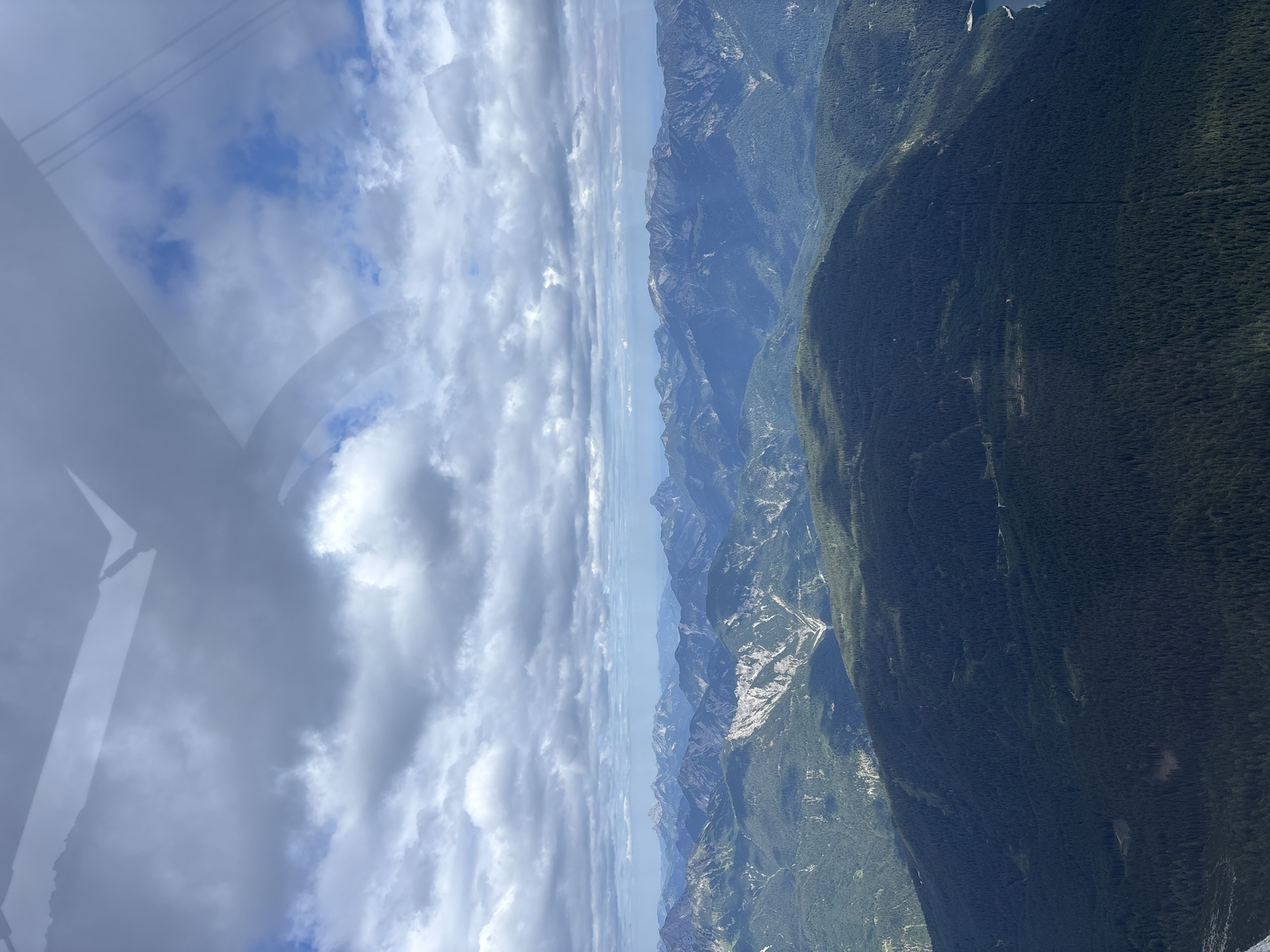 This aerial image captures a panoramic view of the Bavarian Alps, with a focus on the forested slopes and rugged mountain ranges surrounding Grainau, Germany. The foreground is dominated by a dense, dark green coniferous forest covering a large mountain. Beyond this, several layers of mountains recede into the distance, their peaks softened by atmospheric haze. The mountains exhibit varying degrees of vegetation, with some slopes showing patches of grey rock and scree.
The sky is filled with a mix of dramatic, textured clouds. White and grey cumulus clouds are interspersed with patches of bright blue sky, suggesting a dynamic weather system. The sun is not directly visible, but its light casts shadows on the landscape, highlighting the contours of the mountains and forests. A faint reflection, likely from an aircraft window, is visible in the upper part of the image, showing a linear, angular pattern that cuts across the clouds.
While the specific location is identified as being near Grainau, Germany, the image itself does not contain any explicit landmarks or man-made structures that would pinpoint a precise spot within the town or its immediate surroundings. The focus is on the natural, mountainous terrain. There are no people, animals, or active events depicted in the scene; it is a static landscape captured from an elevated perspective.