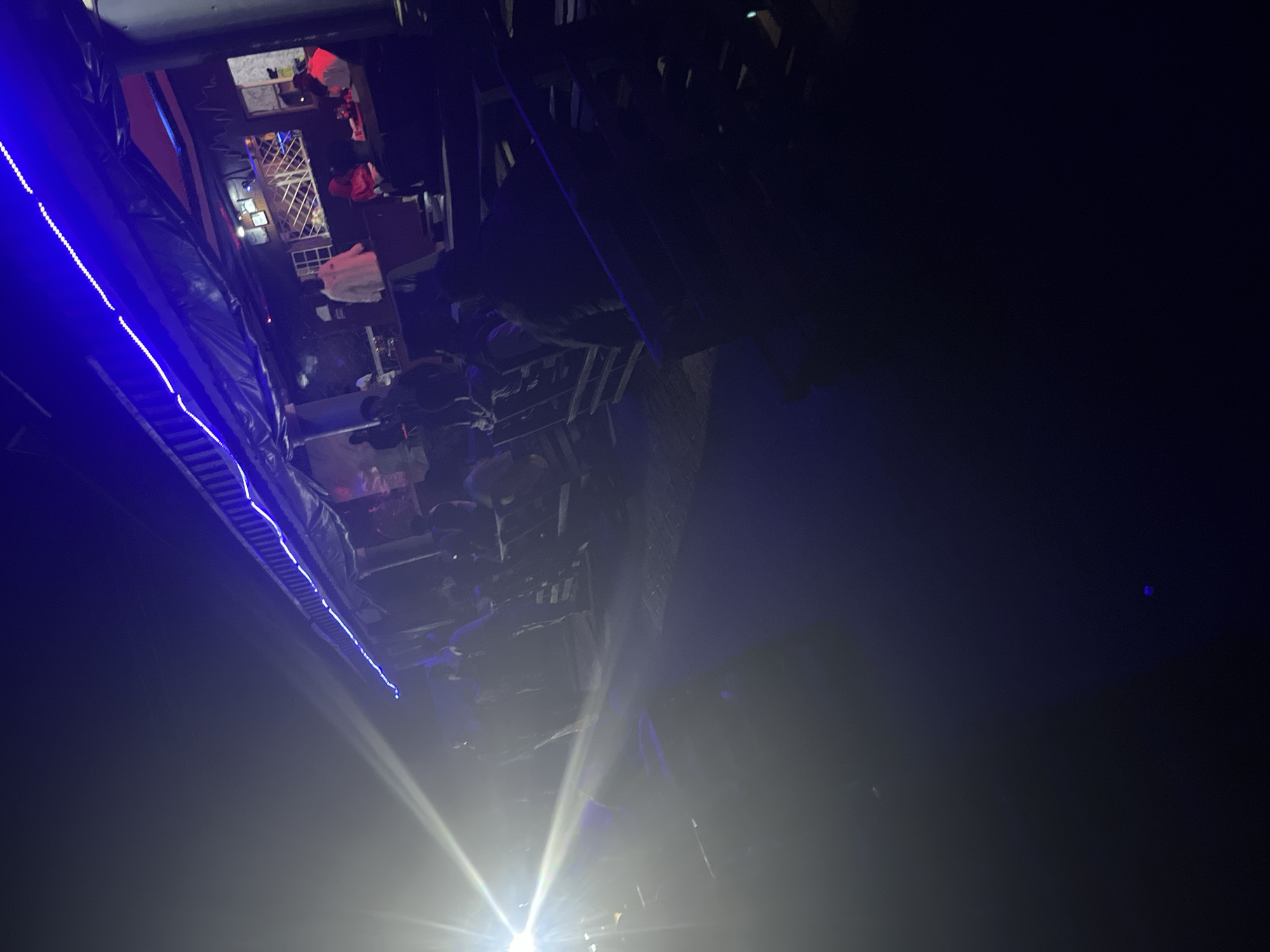 The image depicts an indoor event, likely a party or club, in Durumi, Nigeria. The scene is dimly lit, with prominent blue LED strip lights illuminating a balcony or upper level on the left side of the frame. Below, a crowd of people is visible, though their faces are largely obscured by the low light and a hazy atmosphere, possibly from fog machines or smoke.
Several objects are scattered throughout the space. On the upper level, clothing items, including what appears to be a white hoodie, are draped over railings or surfaces. There are also structures resembling shelves or small stages. In the foreground, a bright light source, possibly a spotlight or flash, creates a strong glare and lens flare, further obscuring details.
The overall mood seems to be energetic and celebratory, consistent with a nighttime entertainment venue. The presence of a crowd suggests an active social gathering. The time of day is certainly nighttime due to the artificial lighting and darkness. There are no visible text elements in the image. The location cues are limited to the context provided, placing the scene in Durumi, Nigeria.