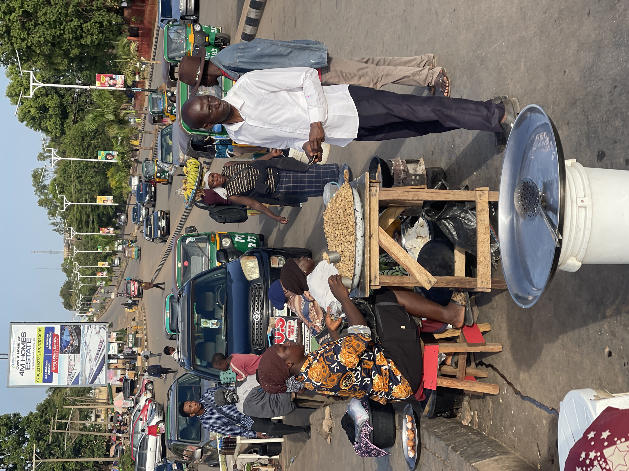 The image depicts a street scene in Jos, Nigeria, where street vendors are selling peanuts to customers. A man in a white shirt stands near a peanut stand, interacting with customers. A woman wearing a patterned top sits near the stand, appearing to be involved in the peanut sale. Other individuals are standing nearby, some possibly purchasing peanuts.
The street is filled with traffic, including cars and commercial tricycles (keke napeps). Advertisements and billboards can be seen in the background.
A detailed breakdown of the image include:
People: A man in a white shirt, a woman in a patterned top, other people buying peanuts, and drivers in the vehicles.
Subjects: Peanut selling activity.
Objects: Peanuts on a stand, wooden stools, cooking utensils, peanuts for sale, cars, keke napeps.
Actions: Selling peanuts, buying peanuts, driving vehicles.
Interactions: Customer-vendor interaction.
Environment: Busy street in Jos, Nigeria, with traffic and commercial activities.
---