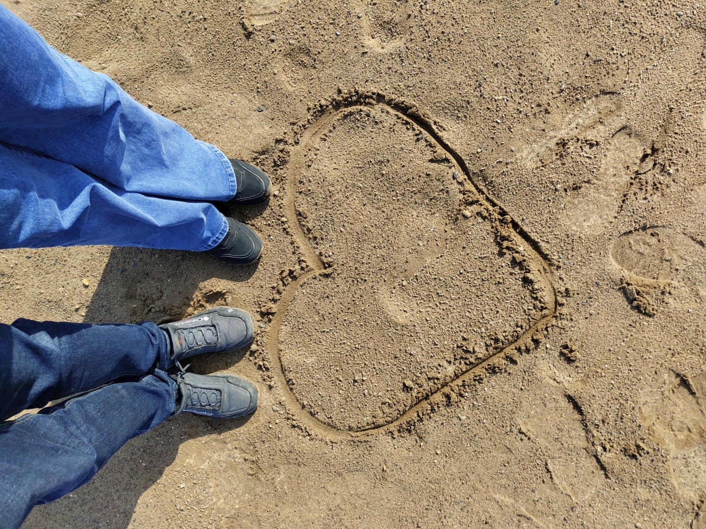 Two people are standing on a sandy surface near a heart drawn in the sand. The person on the left is wearing blue jeans and black shoes. The person on the bottom is wearing dark blue jeans and gray Columbia-branded shoes. The heart is drawn to the right of the two people. The location is not specified.
