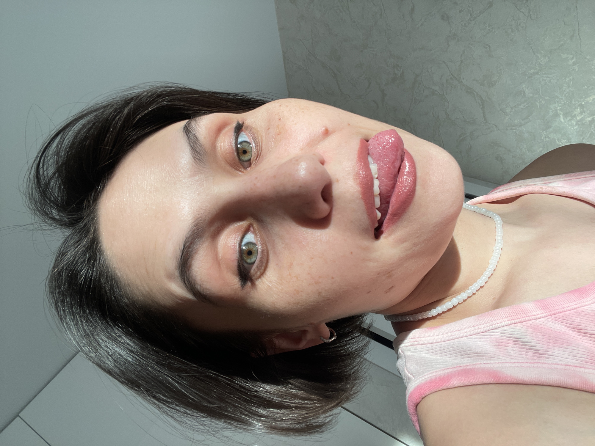 A young woman with short, dark hair is facing the camera and sticking out her tongue. She has green eyes and freckles. She is wearing a pink tie-dye top and a white beaded necklace. The background consists of a textured wall and a white surface. The lighting is bright and appears to be natural sunlight. The image appears to be taken from a low angle, with the camera tilted. There is no information available in this image regarding the city of Izhevsk, Russia.