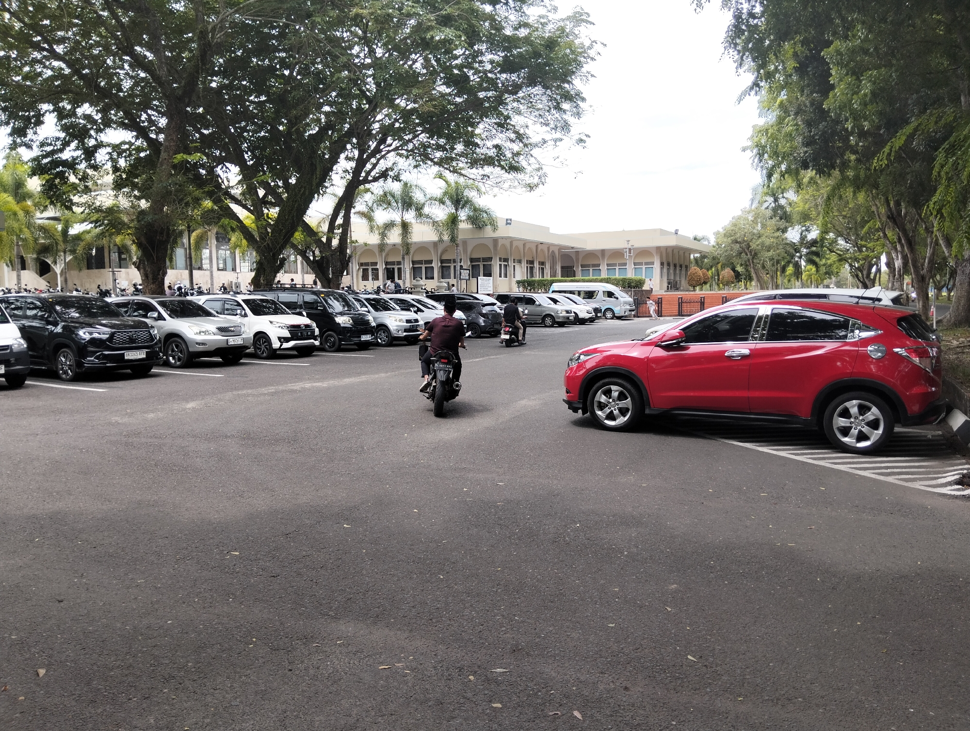 The image depicts an outdoor parking lot in front of a cream-colored building with a covered walkway supported by arches. Several cars, including black SUVs, white sedans, and a red SUV, are parked in rows on an asphalt surface. A motorcycle with a rider is visible in the middle of the parking lot. Lush green trees with dense foliage, some with palm trees interspersed, line the area. The sky is overcast. The scene is likely in Tambontunong, Indonesia, given the architectural style and vegetation typical of the region.