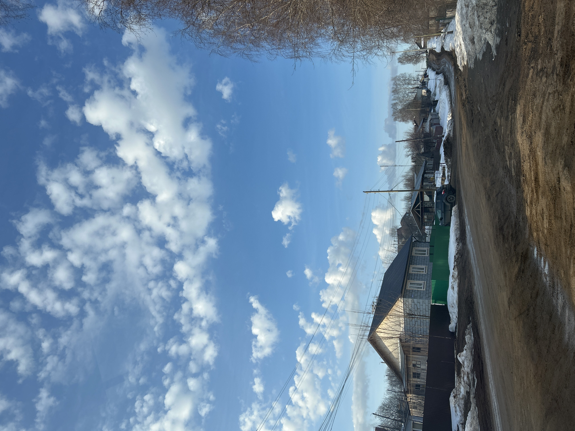 The image depicts a street scene in what appears to be a residential area of Фоки, Russia. The road, muddy and partially covered with melting snow, stretches along the right side of the frame. Adjacent to the road is a row of houses, some with green fencing and dark roofs. A car is parked near one of the houses. Utility poles and wires are visible against the backdrop of a bright blue sky filled with scattered white clouds. Trees with bare branches are visible above the sky.