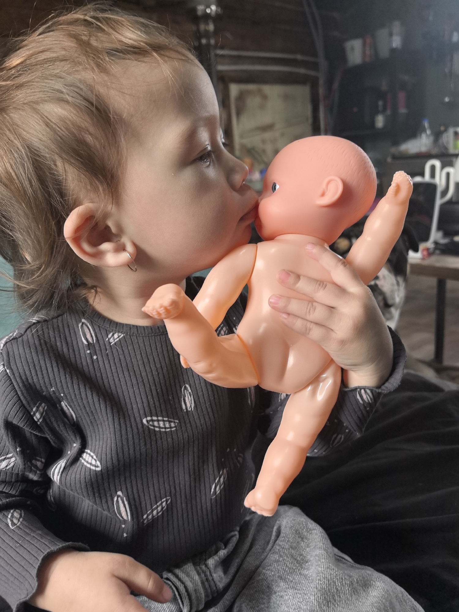 A child is holding and kissing a plastic doll. The child has fair skin and light brown hair, and is wearing a gray, long-sleeved shirt with white patterns. The doll is bald and pink. They appear to be indoors. The location is not discernible from the image.