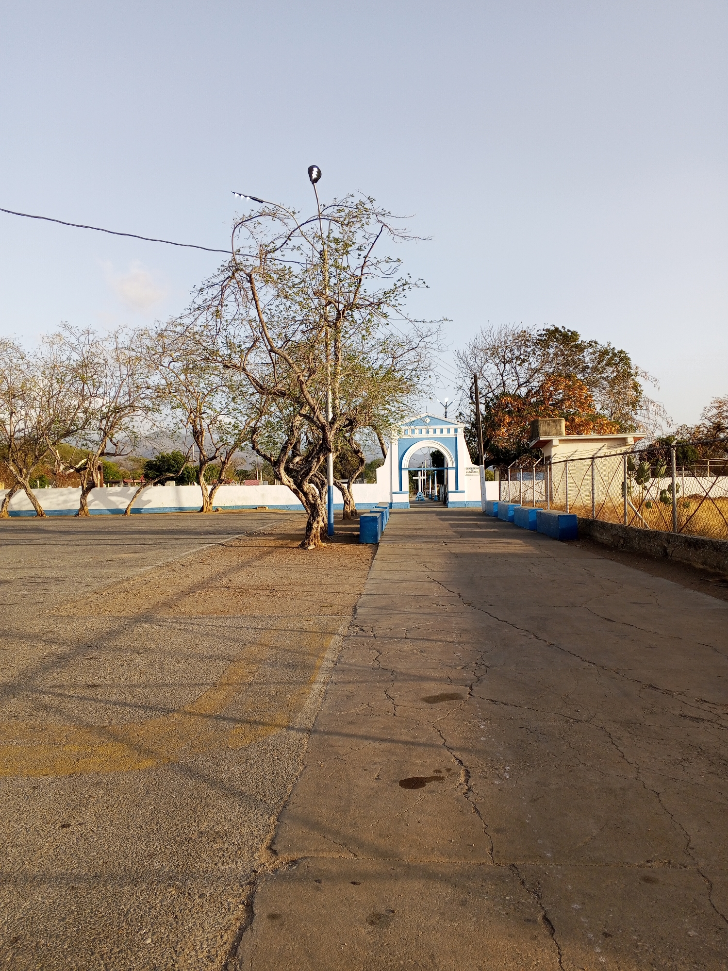 The image shows a street leading to a blue and white arched structure. The street is cracked and worn, with yellow lines painted on the left side. On either side of the street are rows of trees, some of which are bare. There is a white wall with a blue base running along the left side of the street. A chain-link fence runs along the right side of the street, with a small building behind it. There is a light pole in the middle of the trees. The sky is clear and blue. The location is in the city of Unknown, Unknown.