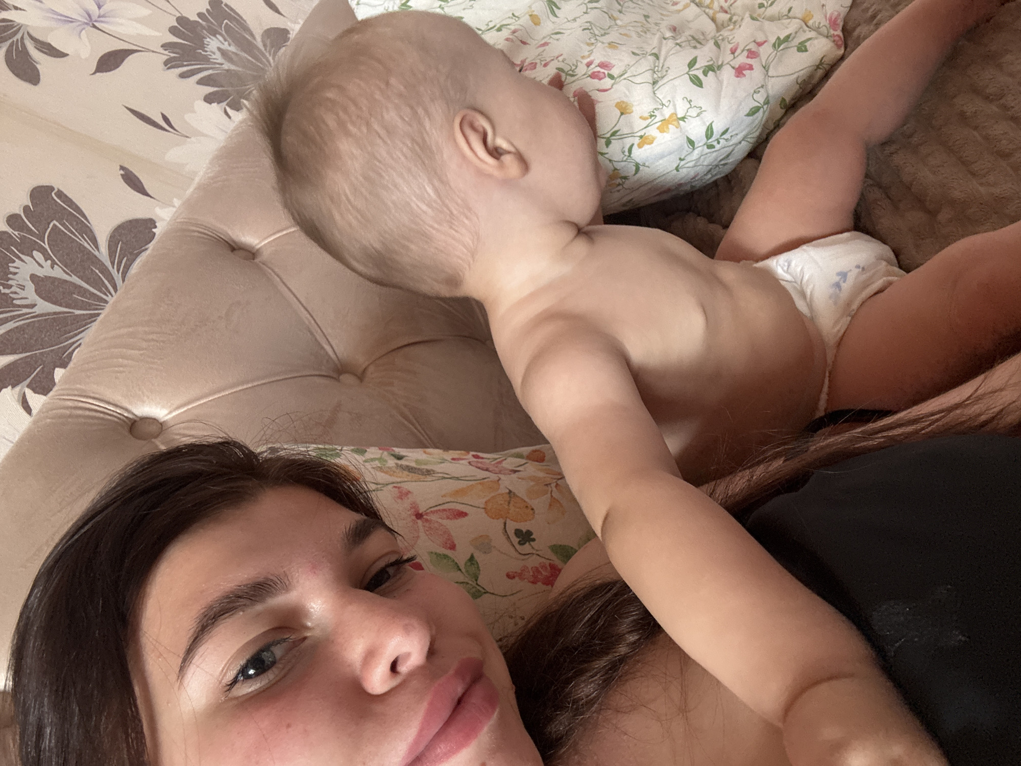 A young woman with dark hair and rosy cheeks is lying in bed, her face close to the camera. Beside her, a baby in a diaper is also lying down, facing away from the camera. The background shows a tufted headboard and patterned wallpaper. The scene appears to be indoors, in a bedroom. The image does not provide any specific details that can be used to determine its location in the city of Klin, Russia.