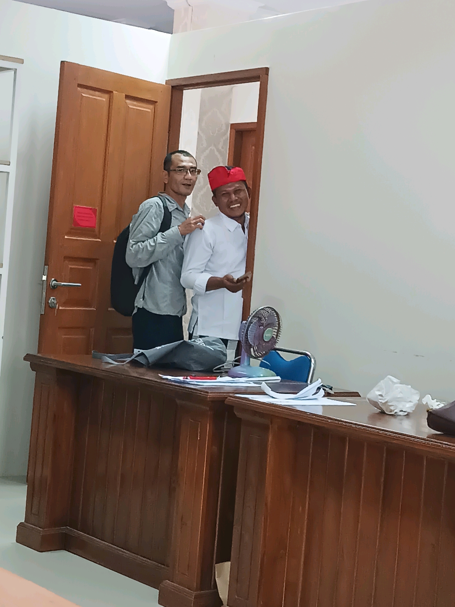 Two men are standing in front of a wooden desk. The man on the left is wearing glasses and a grey shirt. He has a black backpack on his left shoulder. The man on the right is wearing a red hat and a white shirt. He is smiling. Behind the men is a brown door. On the desk, there are papers, a fan, and some crumpled white paper. The scene appears to be indoors, possibly an office.