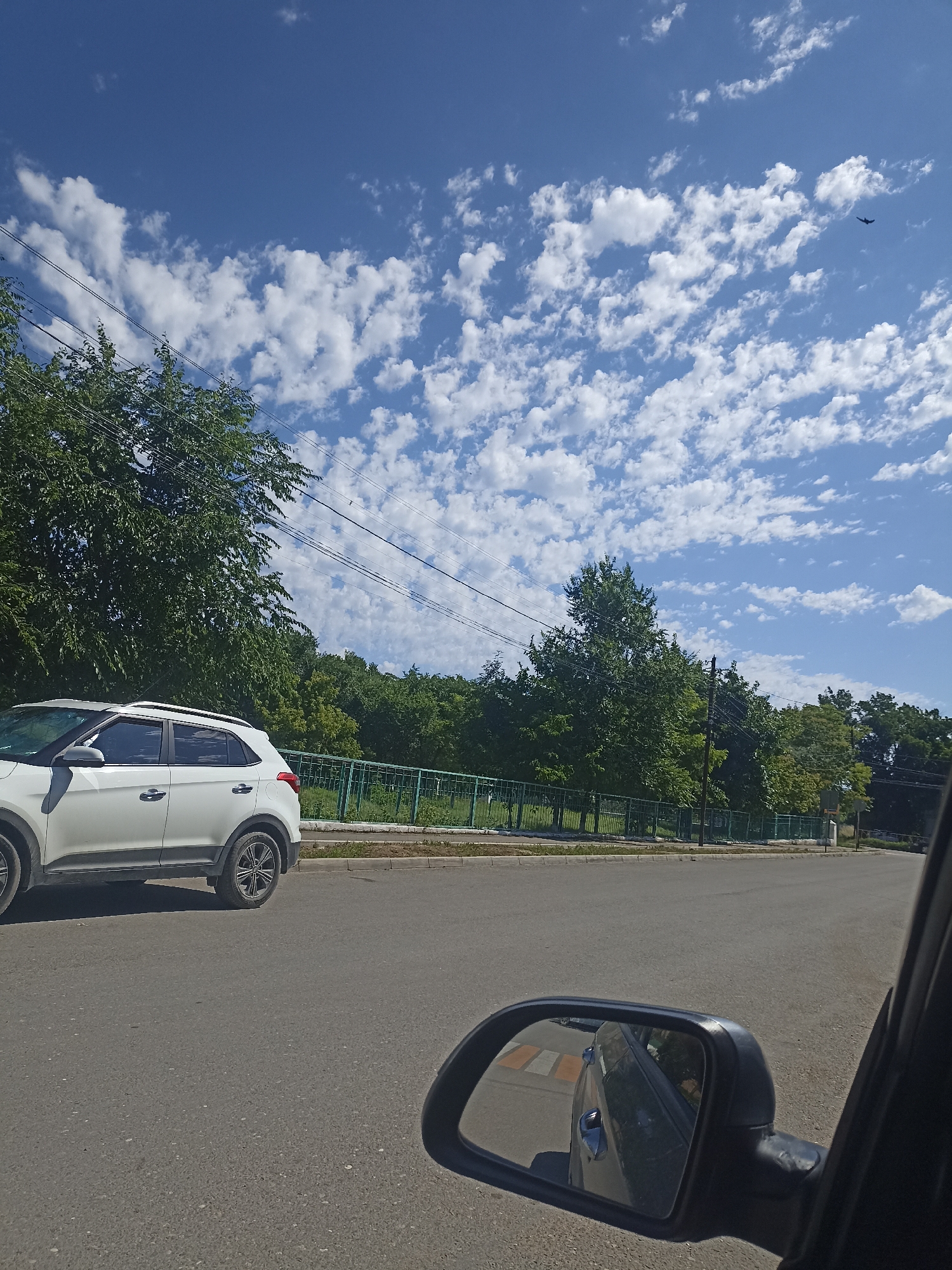 The image shows a view from inside a vehicle, looking out at a street scene. A white SUV is parked on the left side of the road. In the background, there is a line of trees and a green fence. The sky is blue with scattered white clouds. A bird is visible in the sky in the upper right corner of the image. The location is Izberbash, Russia.