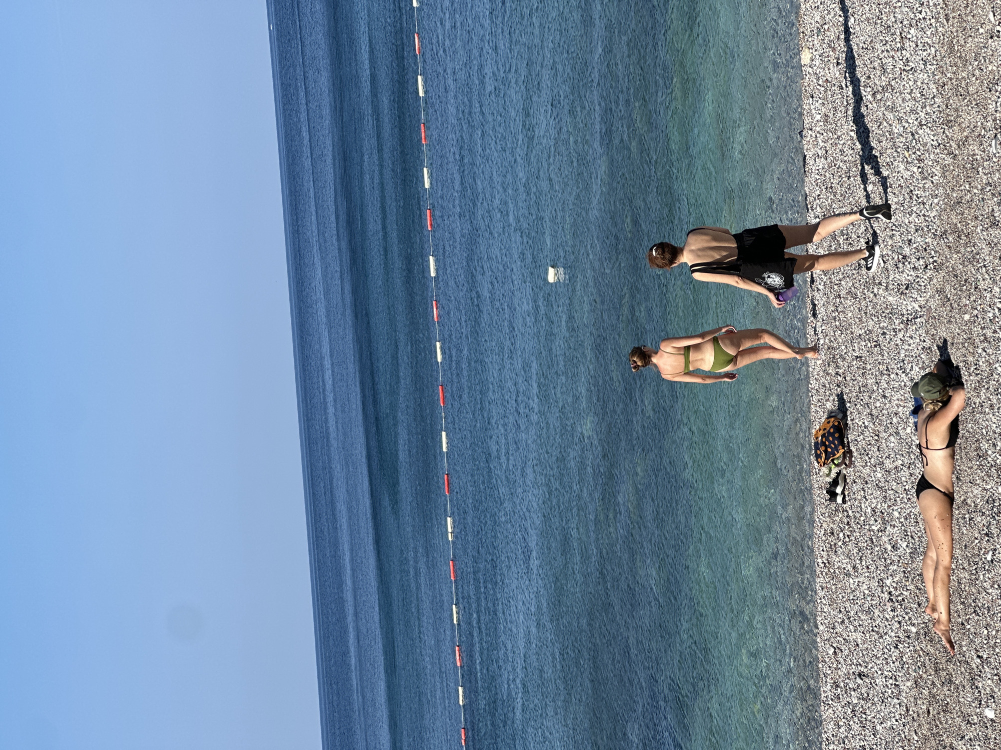 The image shows a sunny day at a pebble beach in Bečići, Montenegro. In the foreground, a person is lying on the beach on their stomach, wearing a black bikini and a green cap. Further back, two individuals are walking towards the clear blue water. One person is wearing a green bikini, and the other is wearing a black swimsuit and carrying a tote bag and a water bottle. A line of buoys marks a boundary in the water. The sea is calm with a gradient of blue hues, and the sky is clear and bright. A patterned beach bag and a pair of black sneakers are placed on the pebbles near the water's edge.
