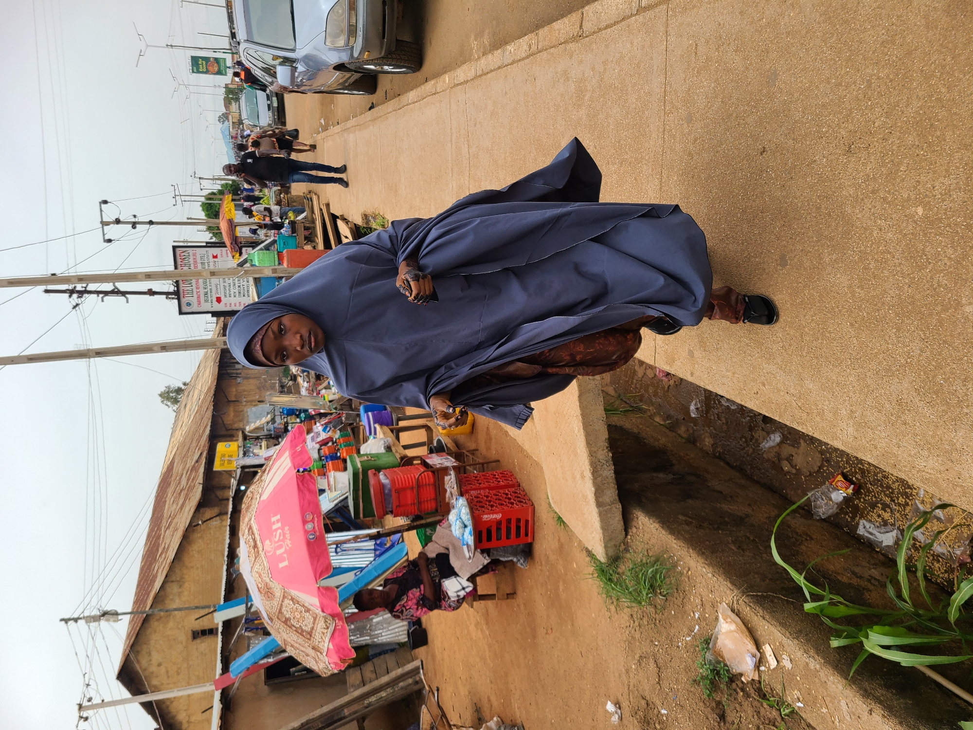 A woman wearing a dark blue hijab and a long robe stands on a sidewalk, looking at the camera. She is holding a dark object in her left hand. In the background, there is a busy market scene with various stalls and people. A silver car is parked on the side of the road. A building with a rusted roof and a pink umbrella bearing the words "LUSH Hair" can be seen. There is trash and foliage in the gutter adjacent to the sidewalk. A sign is visible in the background that reads "THE LOT CHOSEN CHARISMATIC REVIVAL".