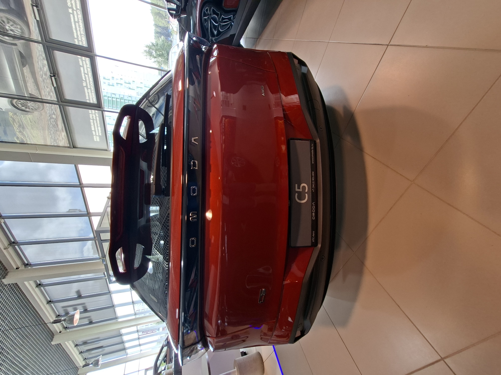 The image shows the rear of a red OMODA C5 car on display inside a dealership. The car has a prominent rear spoiler and the OMODA logo is visible across the tailgate. A license plate frame with the text "C5" is also present. The dealership appears to be modern with large windows and tiled flooring. In the background, other cars and promotional materials are visible. The setting is likely a car showroom in Izhevsk, Russia, given the "OMODA-ITS.ru" website address on the license plate frame, which is a Russian domain. No people are visible in the image.