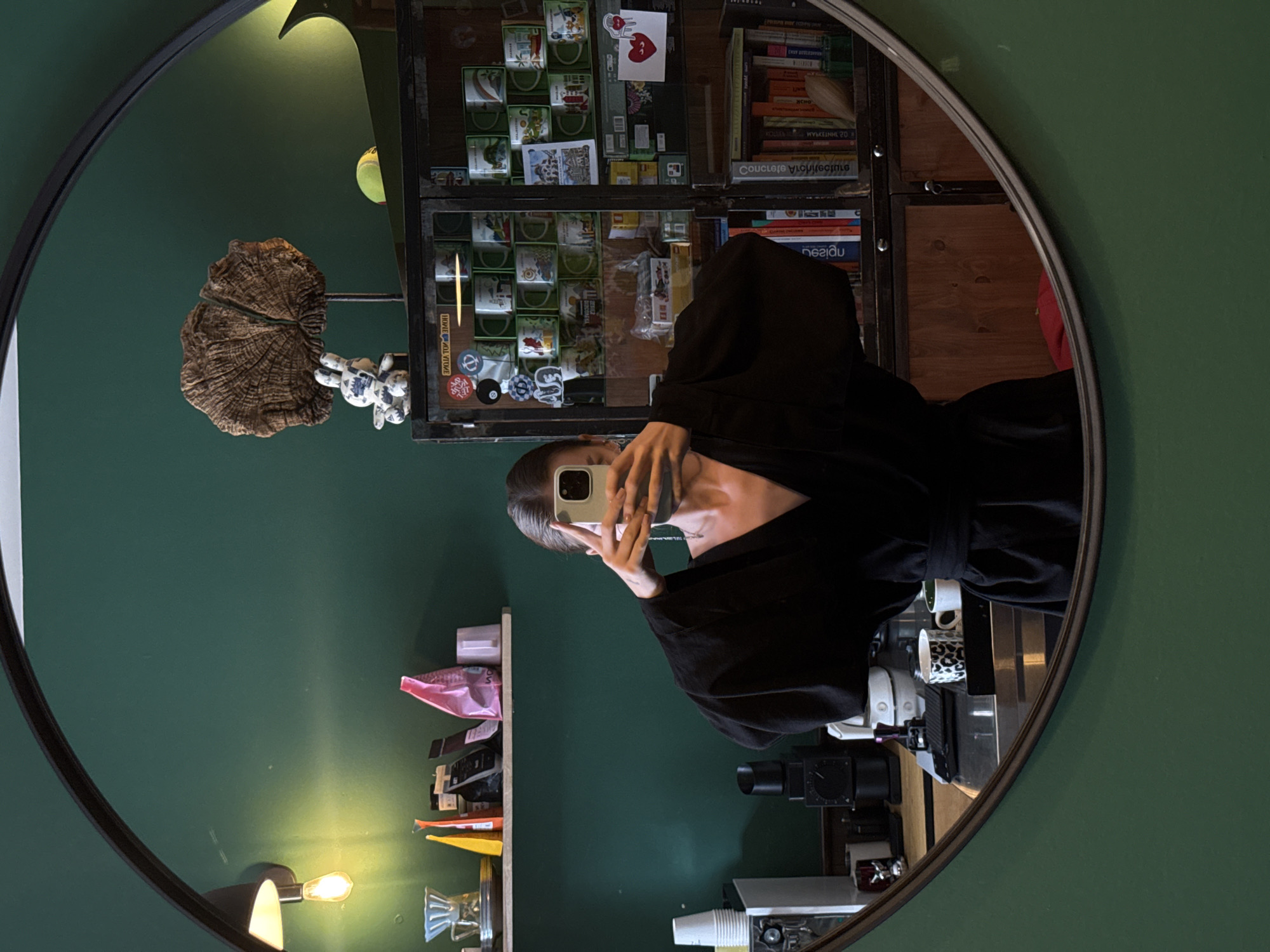 The media file shows a reflection in a circular mirror. The main subject is a person taking a selfie, wearing a black robe or cloak. They are holding a smartphone in their right hand, with their left hand partially visible. The person has dark hair and is looking at the camera.
The background of the reflection reveals a room with a dark green wall. On the left side of the reflection, there are shelves with various items, including what appears to be a decorative object resembling a dried plant or fungus, and a small white stuffed toy. Further down the shelves, there are bags and packaged items.
On the right side of the reflection, there is a glass-fronted cabinet filled with what look like small boxes or packages, possibly containing mugs or collectibles. Above and to the right of this cabinet are books on shelves. The shelves also contain more books and some decorative items. A light fixture with a yellow bulb is visible on the left side of the reflection, casting a warm glow.
The overall scene appears to be an indoor setting, likely a room in a home or a shop. There are no clear indications of the time of day or weather conditions. The mood seems casual and personal, typical of a selfie.
Visible text in the reflection includes titles on book spines, but they are mostly unreadable due to the reflection and image quality. There are also some small stickers or labels on the glass cabinet, some of which appear to have Cyrillic lettering, which is consistent with the location context of Odesa, Ukraine. Some of these stickers have circular designs or logos. The text on the books is too blurred to decipher.