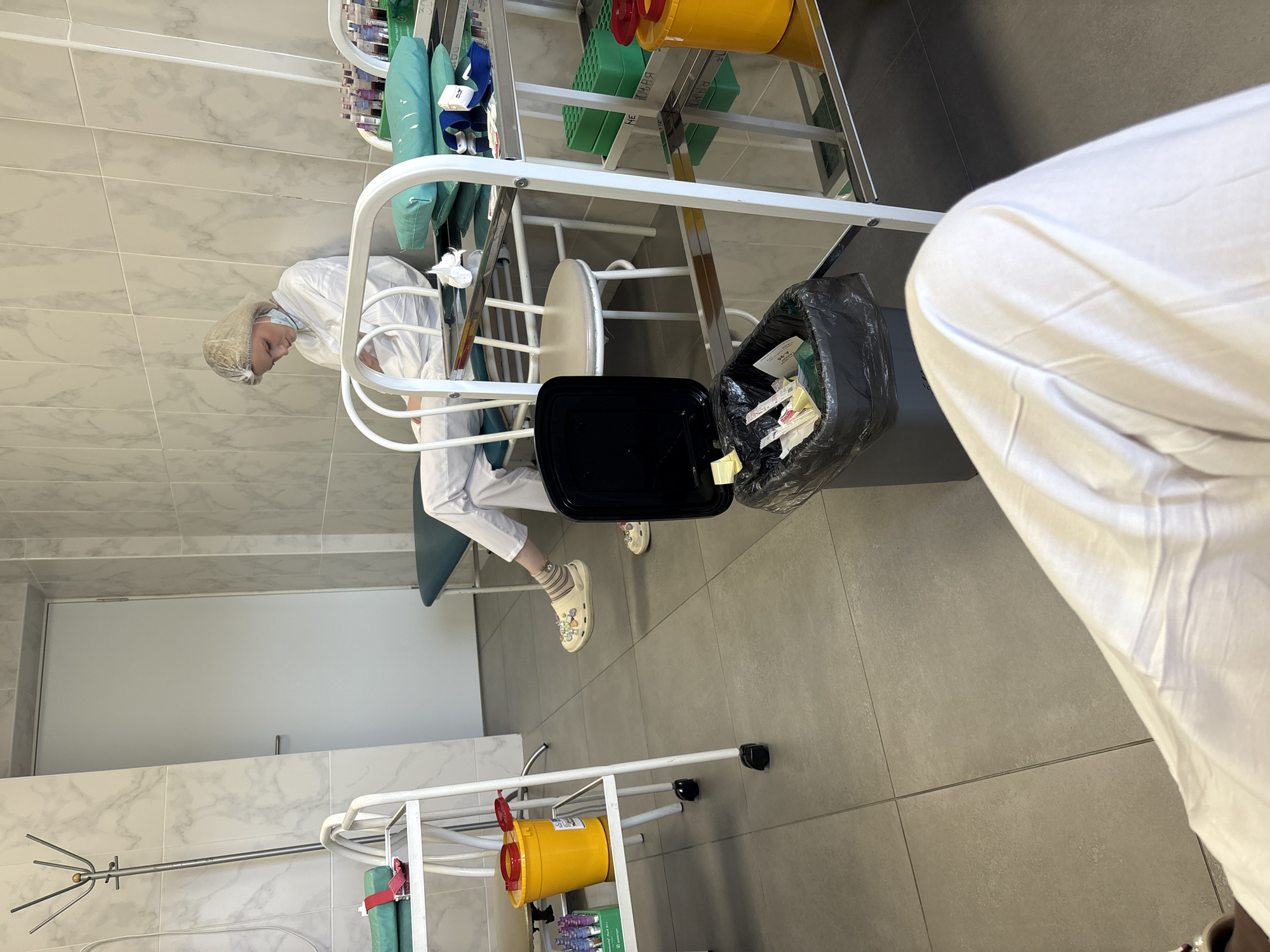 A woman in a white lab coat and hairnet sits on a medical examination table in a room with tile walls. She leans forward slightly, her face partially obscured. Next to her is a small black trash can with a black plastic bag inside, containing what appears to be used medical supplies and paper products. In the background, there is a white metal shelf unit with various medical supplies and equipment. Also visible are yellow biohazard containers and a closed white door. Based on the interior design and medical equipment, this is most likely a medical office or examination room, though the exact location within Saint Petersburg, Russia, is impossible to determine from the image alone.