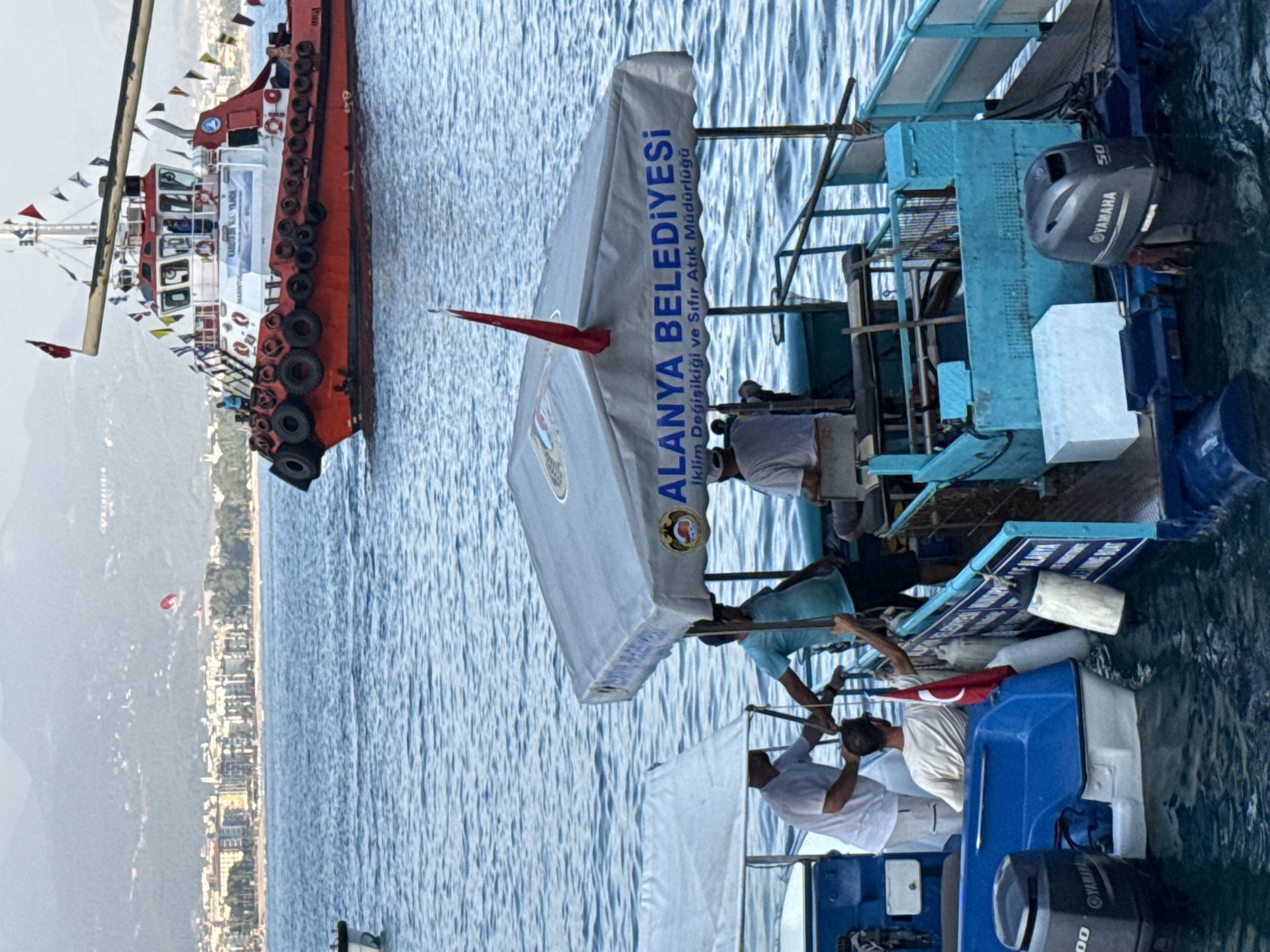 This media file shows a view from a boat on the water, likely in Alanya, Turkey. The main focus is on two boats. The boat in the foreground is a blue and white vessel with several people on board. One person is operating a Yamaha outboard motor, identified by the "YAMAHA 50" text. Another person is holding a Turkish flag. A large white canopy is also visible on this boat, with text in Turkish that reads "ALANYA BELEDİYESİ İklim Değişikiği ve Sıfır Atık Müdürlüğü", which translates to "Alanya Municipality Climate Change and Zero Waste Directorate". This suggests the boat might be associated with a municipal service.
In the background, a larger red and white vessel is partially visible, appearing to be a tugboat or similar workboat. It is adorned with numerous small flags. The sea is calm and blue, and the distant shoreline shows buildings and hills, typical of a coastal city. The lighting suggests it is daytime, possibly late morning or early afternoon. The overall scene appears to be a calm day on the water, with a sense of activity related to boating and perhaps municipal duties.