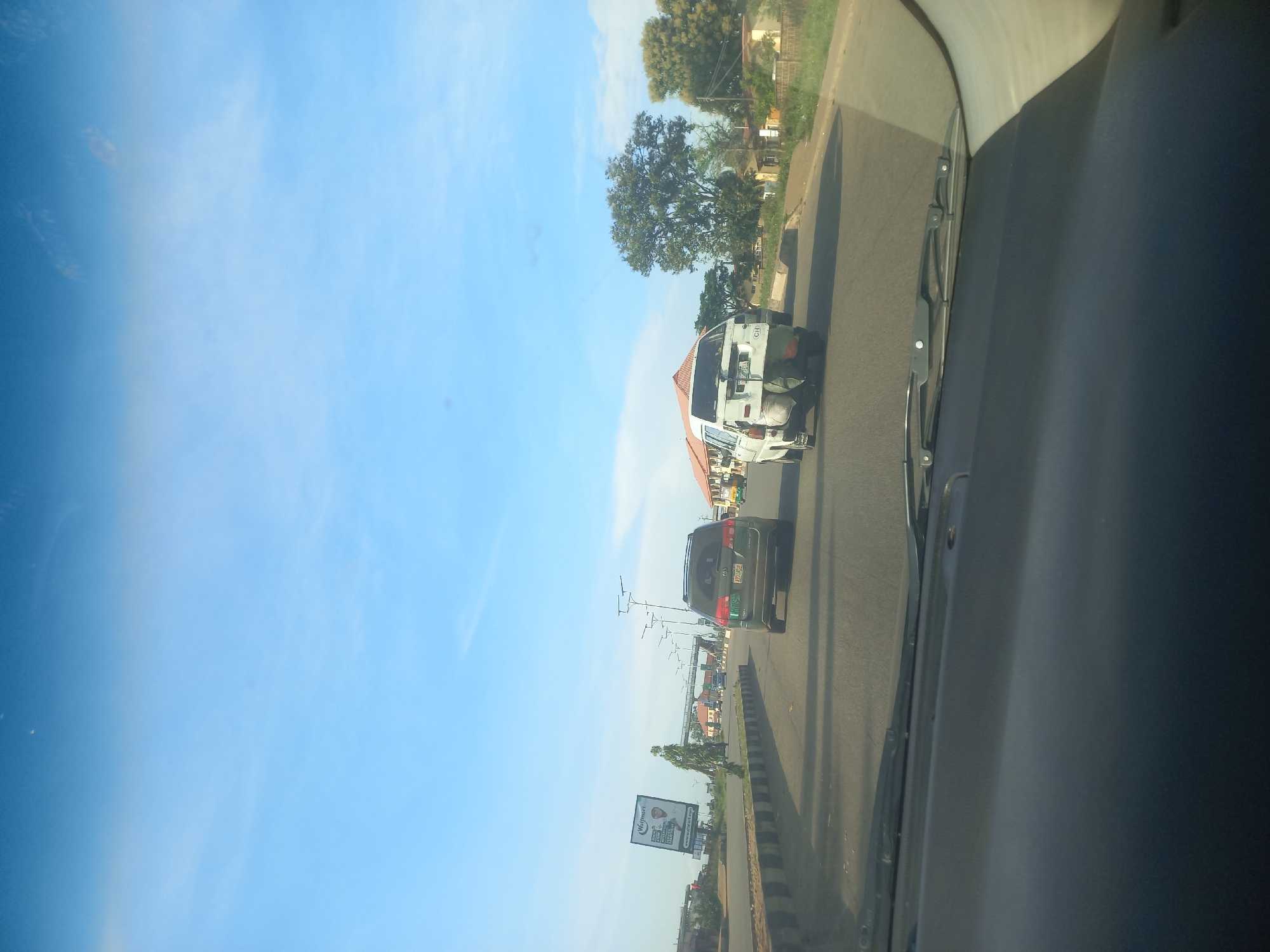 A clear, daytime, eye-level shot captures a view from inside a vehicle on a highway. The sky is a bright, cloud-streaked blue. In the foreground, the dark grey dashboard and windshield wipers of the vehicle are visible. The highway itself is asphalt, with a concrete barrier separating lanes.
Several vehicles are on the road. A dark-colored SUV is in the right lane, followed by a white van. The van appears to be carrying luggage or cargo tied to its rear. Further down the road, other cars and trucks are present, with some buildings and trees lining the sides of the highway. A billboard advertising "Waffmart" is visible on the left side of the road, with a pedestrian bridge overhead. The scene suggests a typical day on a moderately busy highway.