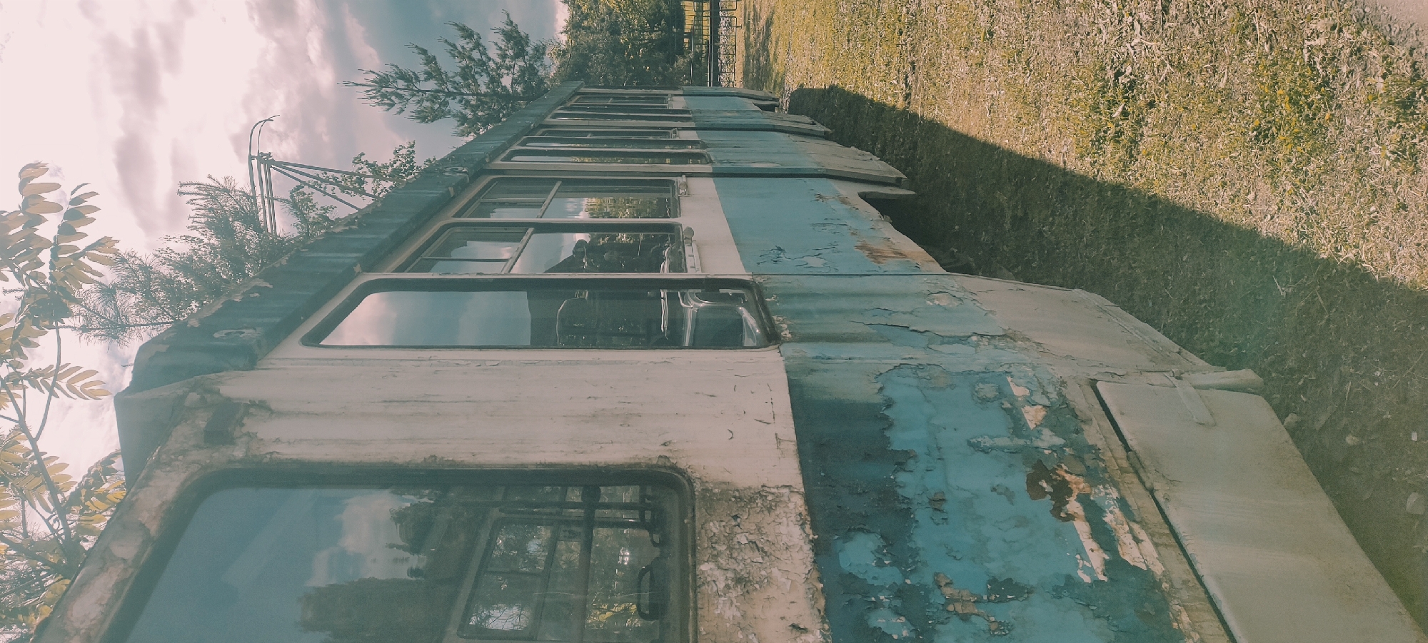 The image shows a weathered, derelict train car situated in a grassy area. The train car is primarily blue and white, with significant paint peeling and rusting. Multiple windows are visible, some reflecting the surrounding environment. The train car is partially in the shade, suggesting a nearby structure or tree. The sky above is cloudy. The photograph may be taken in the city of Ivanovskoe, Russia.