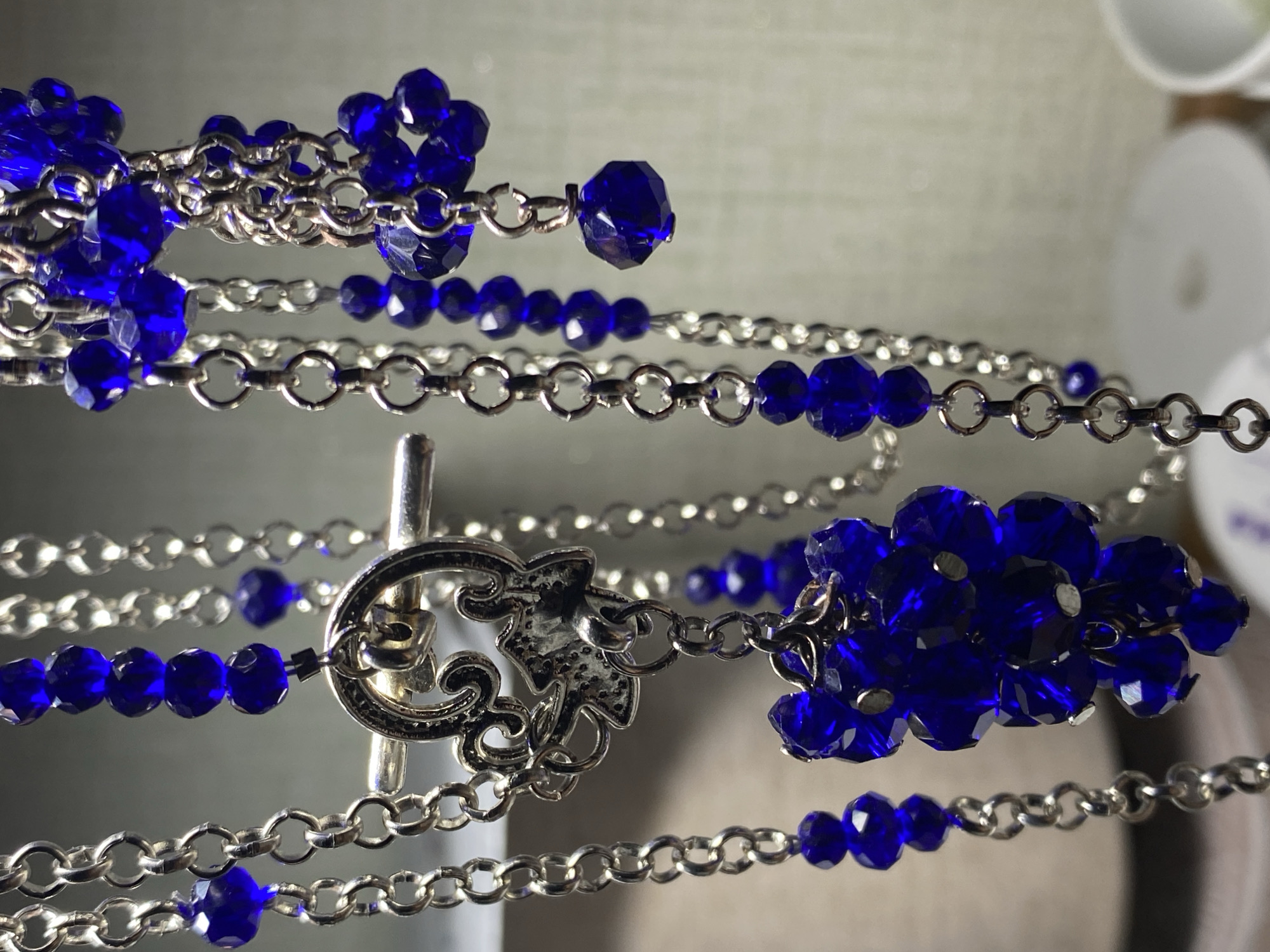A close-up shot focuses on multiple silver chains adorned with faceted blue beads and charms. One prominent charm appears to be a silver toggle clasp with an ornate design, possibly depicting an animal. The chains hang vertically against a blurred background of neutral tones, suggesting an indoor setting, potentially a craft or personal space. There are no people or discernable elements of a city environment present in the image. The lighting is diffused, highlighting the reflective surfaces of the metal and the sparkle of the beads. The scene is static, with no actions or interactions occurring.