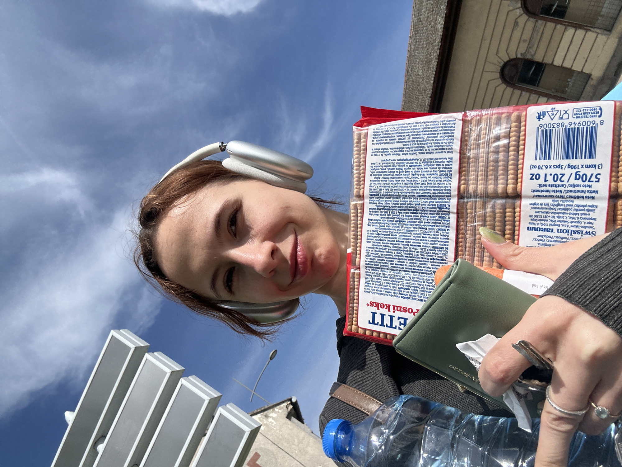 A young woman is featured in a close-up, upward-angled selfie, smiling subtly towards the camera. She wears large, silver over-ear headphones and a dark long-sleeved top, with her reddish-brown hair framing her face. She has light green nail polish on her right hand.
In her right hand, she holds a package of "ETIT Posni keks" (biscuits) by the brand "Swisslion Takovo," with the weight clearly displayed as "570g / 20.1 oz." The packaging also lists "Republika Srbija" as the country of origin. Her left hand holds a green wallet, embossed with "Pellezzo," and a set of keys. A plastic water bottle with a blue cap is visible below her hand. The wallet contains a slip of paper with "Novi Sad" printed on it, indicating the location.
The scene is outdoors on a bright day, dominated by a clear blue sky with scattered light clouds. Parts of an old building with arched windows are visible in the upper right, and modern architectural elements or signage are in the lower left, suggesting an urban setting. The overall mood is cheerful and relaxed.
