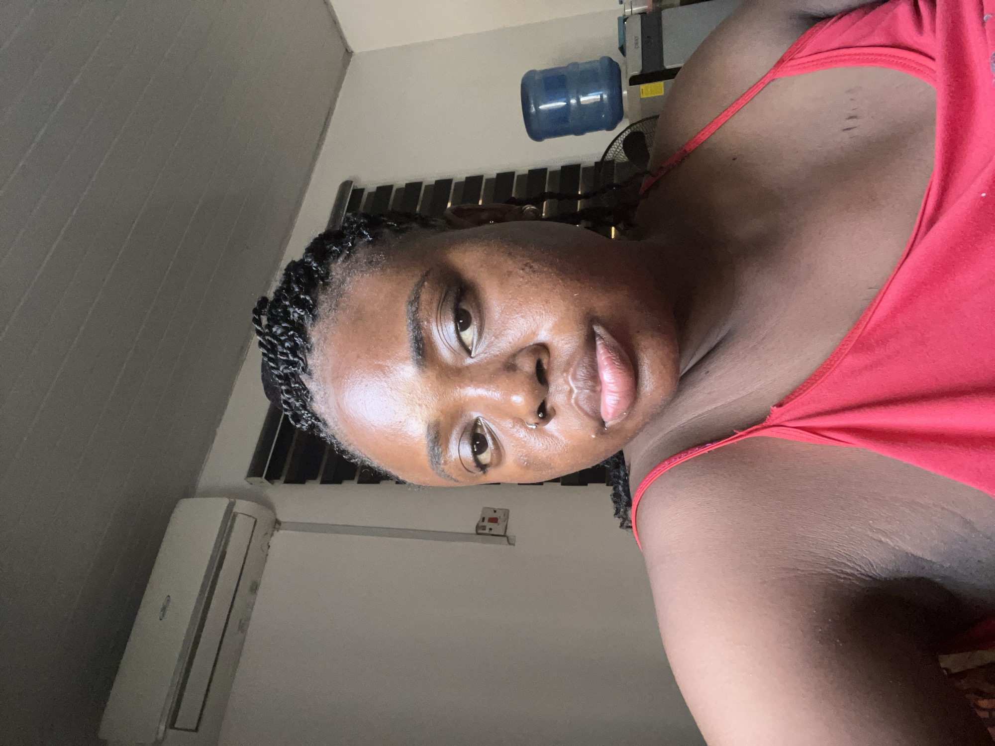 The image shows a close-up, head-and-shoulders portrait of a young Black woman with braided hair. She is wearing a red tank top and looking directly at the camera with a neutral expression. Her skin appears oily and has some minor blemishes. She has a nose ring and a visible birthmark on her chest.
In the background, an air conditioning unit is mounted high on a white wall. To the right, a blue water cooler jug sits on top of a black appliance. Behind the woman, a window with horizontal blinds is visible. The overall setting appears to be an indoor room, possibly a bedroom or living space, in Abuja, Nigeria.