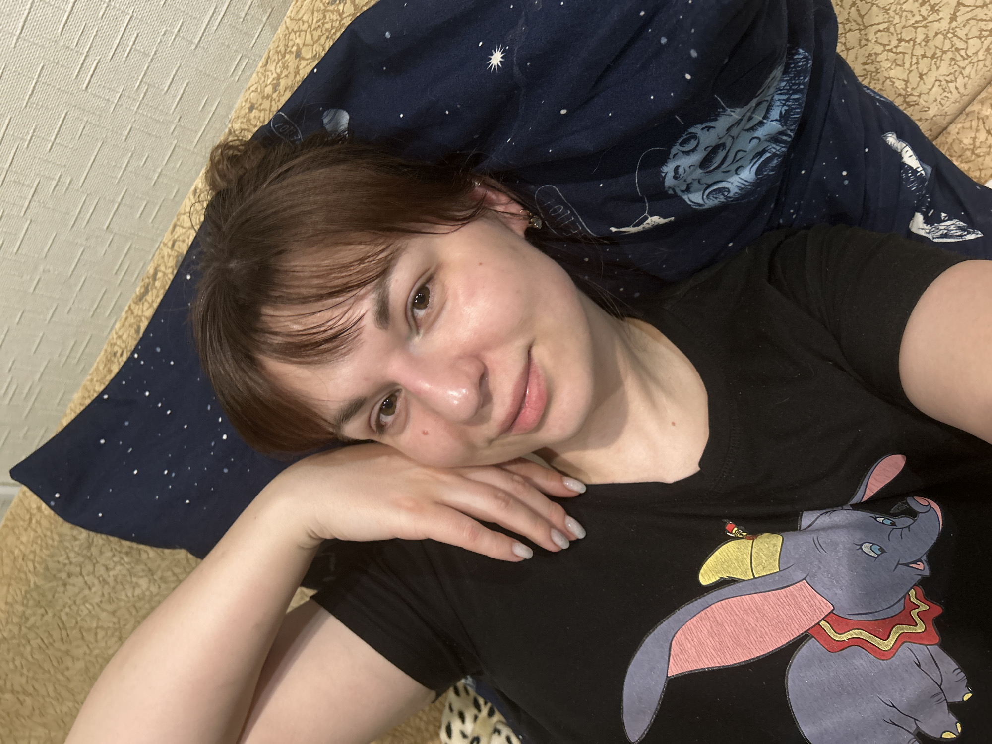 A young woman lies on a bed with a galaxy-patterned pillow behind her head. She is wearing a black T-shirt with a Dumbo print on it. Her brown hair is partially up in a bun. The bed has a textured, tan cover. It is not possible to identify the location in Фоки, Russia, based on this image.