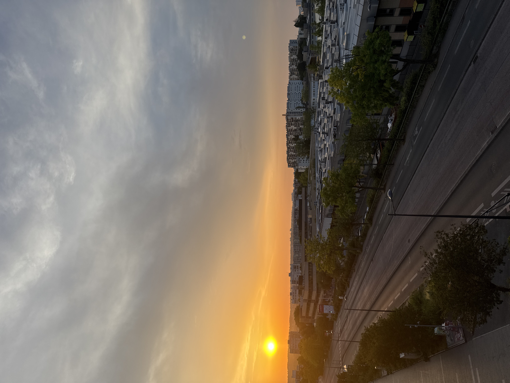 The image depicts a city skyline at sunset. The sun, a bright orange orb, is low in the sky on the left side of the frame, casting a warm glow over the scene. The sky transitions from a vibrant orange near the horizon to a pale gray with scattered clouds towards the top.
In the foreground, a multi-lane road runs diagonally from the bottom left to the middle right of the image. The road appears to be asphalt with white lane markings. A sidewalk runs along the left side of the road, with some trees and streetlights.
Beyond the road, there are various buildings, including a long, low-rise industrial or commercial complex with a flat, silver-colored roof and numerous ventilation units. Further back, a cluster of taller residential buildings with multiple stories and balconies are visible, appearing light-colored. Trees with green foliage are scattered throughout the scene, softening the urban landscape.
The overall impression is an urban environment during golden hour, with the city bathed in the soft light of the setting sun. The scene is quiet, with no people or vehicles clearly visible.