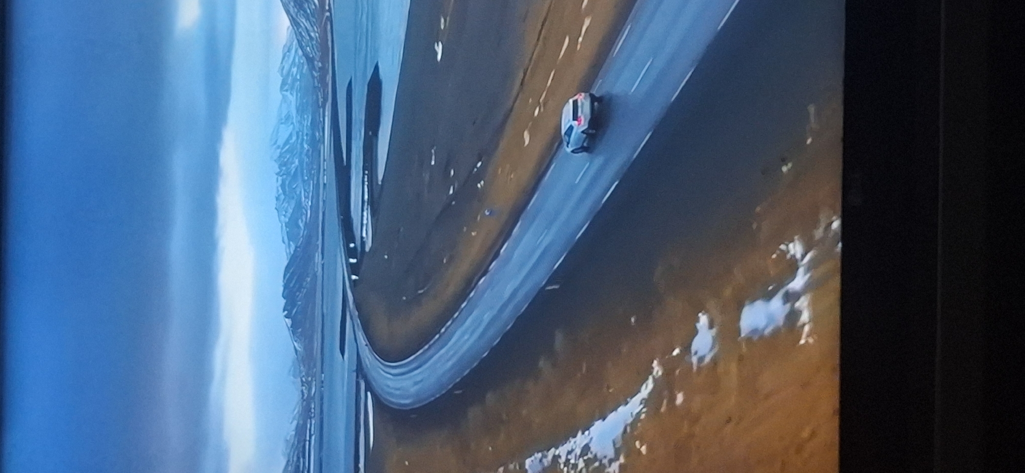 The image depicts an aerial view of a road curving through a landscape of water, dark land, and distant snow-capped mountains under a cloudy blue sky. A white SUV is driving on the right side of the wet road. The foreground shows patches of snow on brown, dry ground. There is no indication that this scene is located in Bila Tserkva, Ukraine, as there are no urban elements or identifiable landmarks suggestive of that location.