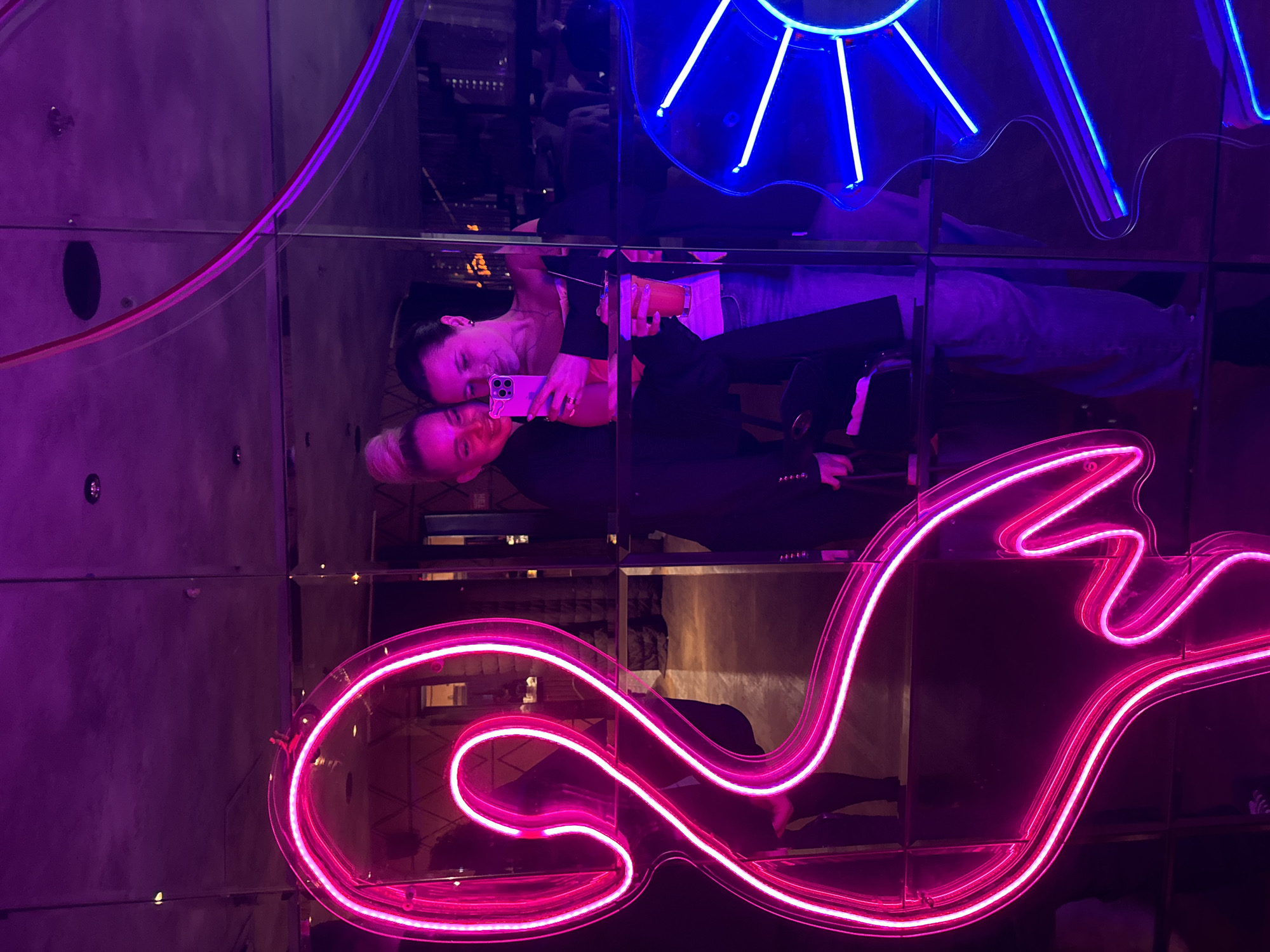 A vertical shot shows two women in a mirrored environment, with neon lights in the background. One woman, with short blonde hair, is in the foreground, smiling and holding a pink iPhone. The other woman, with dark hair pulled back, is slightly behind, also smiling and holding a cup. They appear to be taking a selfie. A blue neon light fixture, resembling a sun or star, is in the upper portion of the shot, and a larger pink neon light, shaped like a stylized wave or script, dominates the lower portion. The mirrored walls reflect the women and neon lights, creating a layered and distorted effect. The location is Krasnogorsk, Russia.