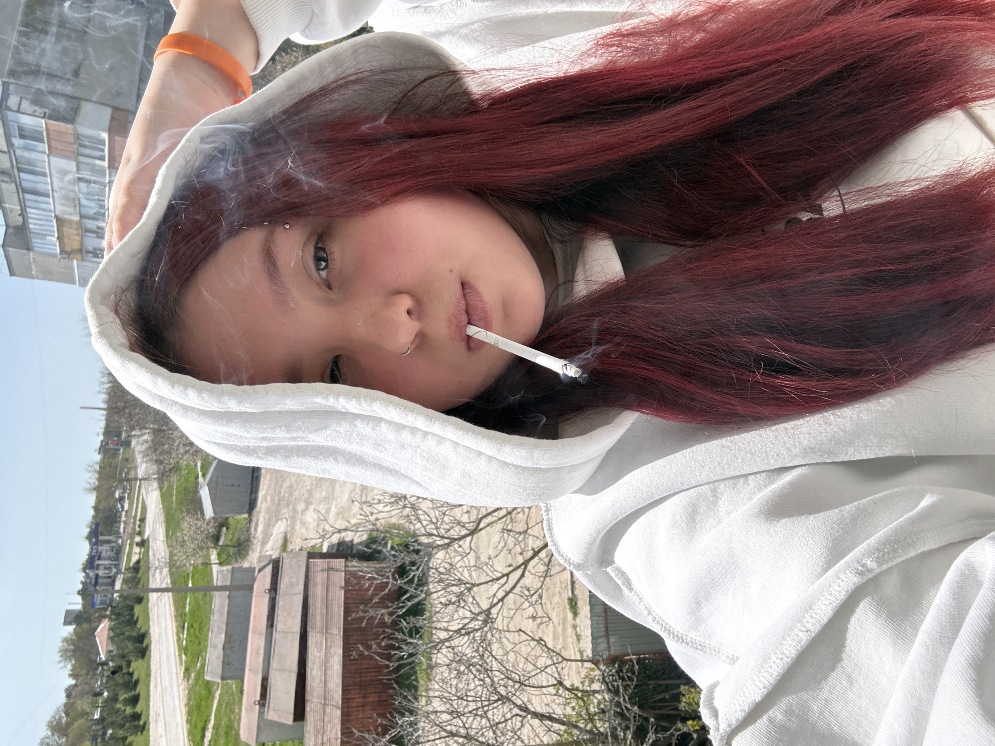 A young person with reddish-brown hair, a nose ring, and a possible eyebrow piercing is shown wearing a white hooded sweatshirt with the hood up. They are holding a lit cigarette in their mouth, with smoke visible around the cigarette. The background includes buildings and greenery, suggesting an urban or suburban location. This image was taken in Chornomorskoe, Ukraine.