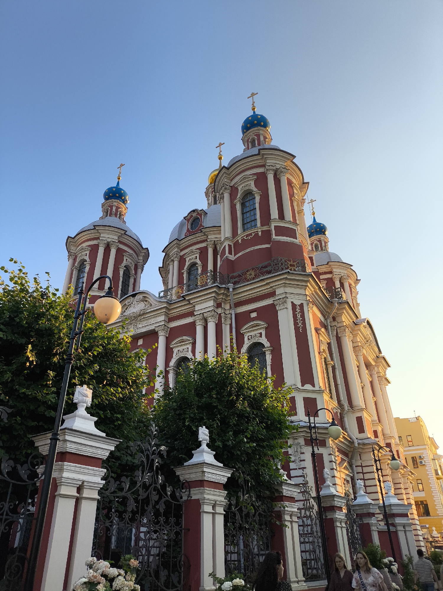 A low-angle shot captures a vibrant red church with white and gold detailing against a clear, pale blue sky. The church features multiple ornate domes, the largest ones adorned with blue and gold stars, and topped with crosses. The architecture is elaborate, with columns, arched windows, and decorative fencing.
In the foreground, lush green trees partially obscure a black wrought-iron fence and decorative stone pillars. A street lamp with a glowing, pale yellow bulb is visible on the left. Several people are walking along the street in front of the fence, though their faces are mostly obscured by the angle and foliage. One person in the center appears to be looking towards the camera.
The lighting suggests it is either early morning or late afternoon, with a warm glow illuminating the side of the church. The sky is clear, indicating fair weather. There is no visible text in the image. The overall scene conveys a sense of architectural beauty and a peaceful, possibly urban, setting.